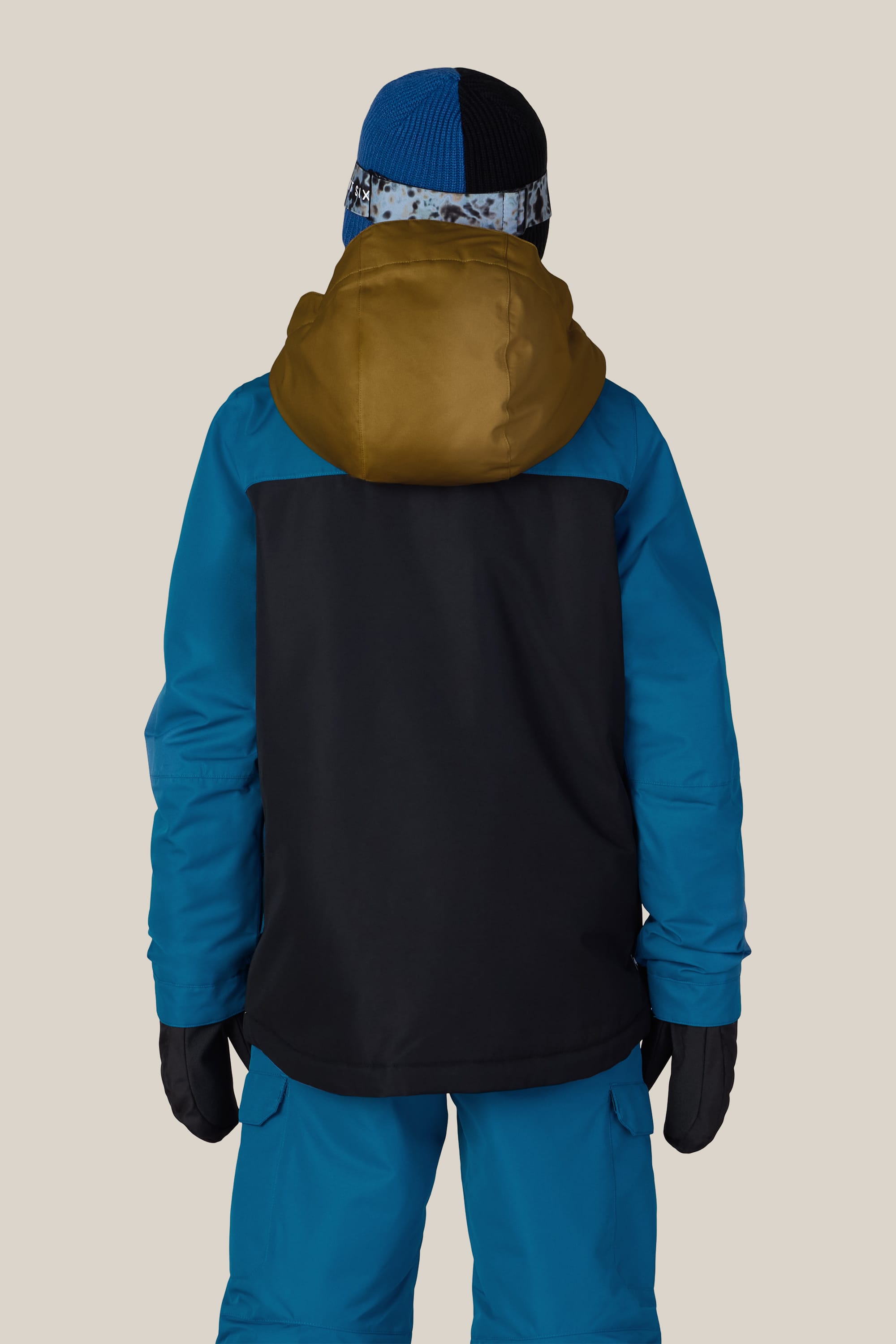A child is seen from the back against a plain light background wearing the 686 Boys’ Gateway™ Insulated Jacket by 686 in blue and black with a brown hood, paired with matching blue pants, black gloves, and a blue knit hat.