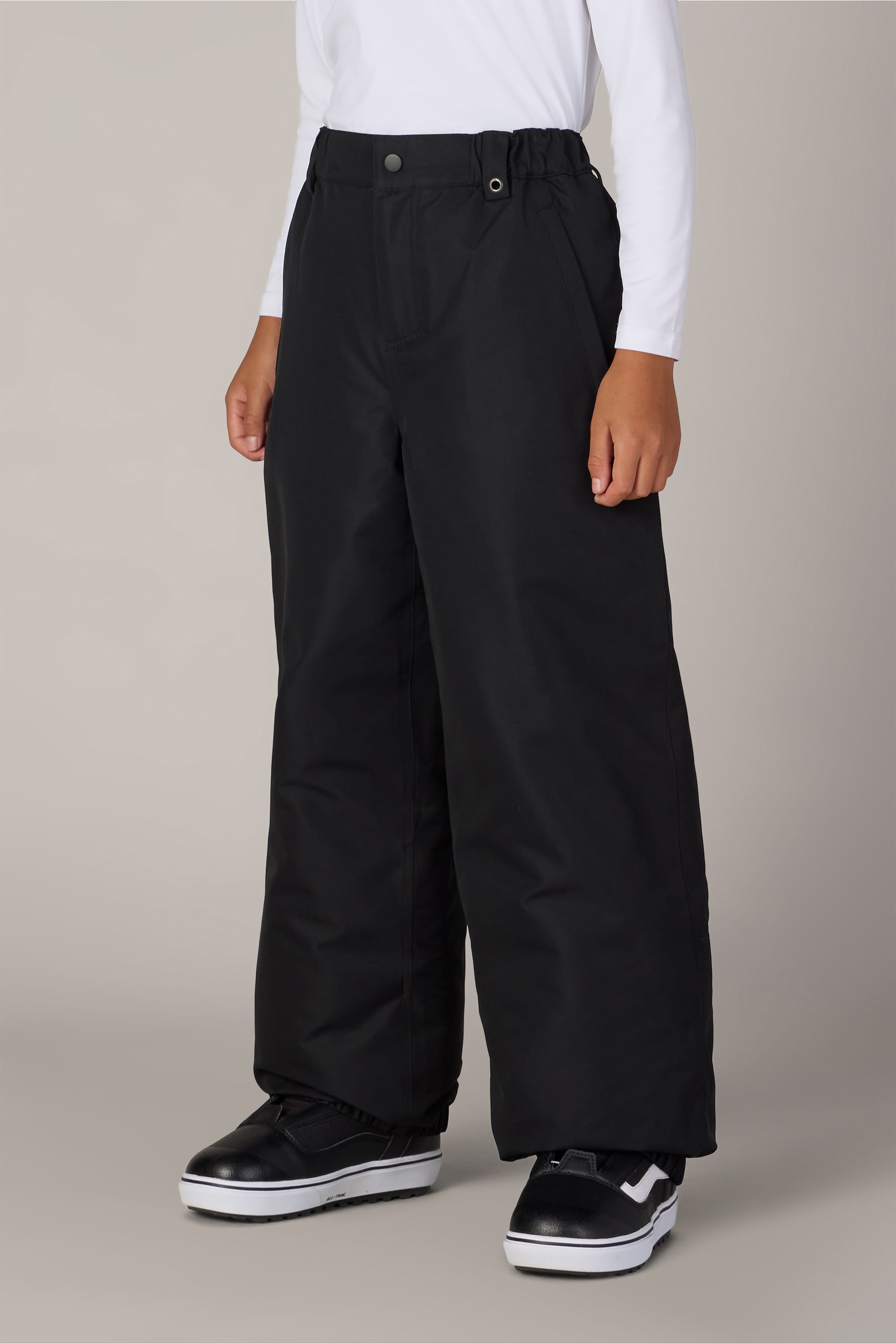 A child wearing a white long-sleeve shirt, black 686 Boys’ 686 Dojo® Insulated Pants from 686, and black-and-white sneakers stands against a plain light background.