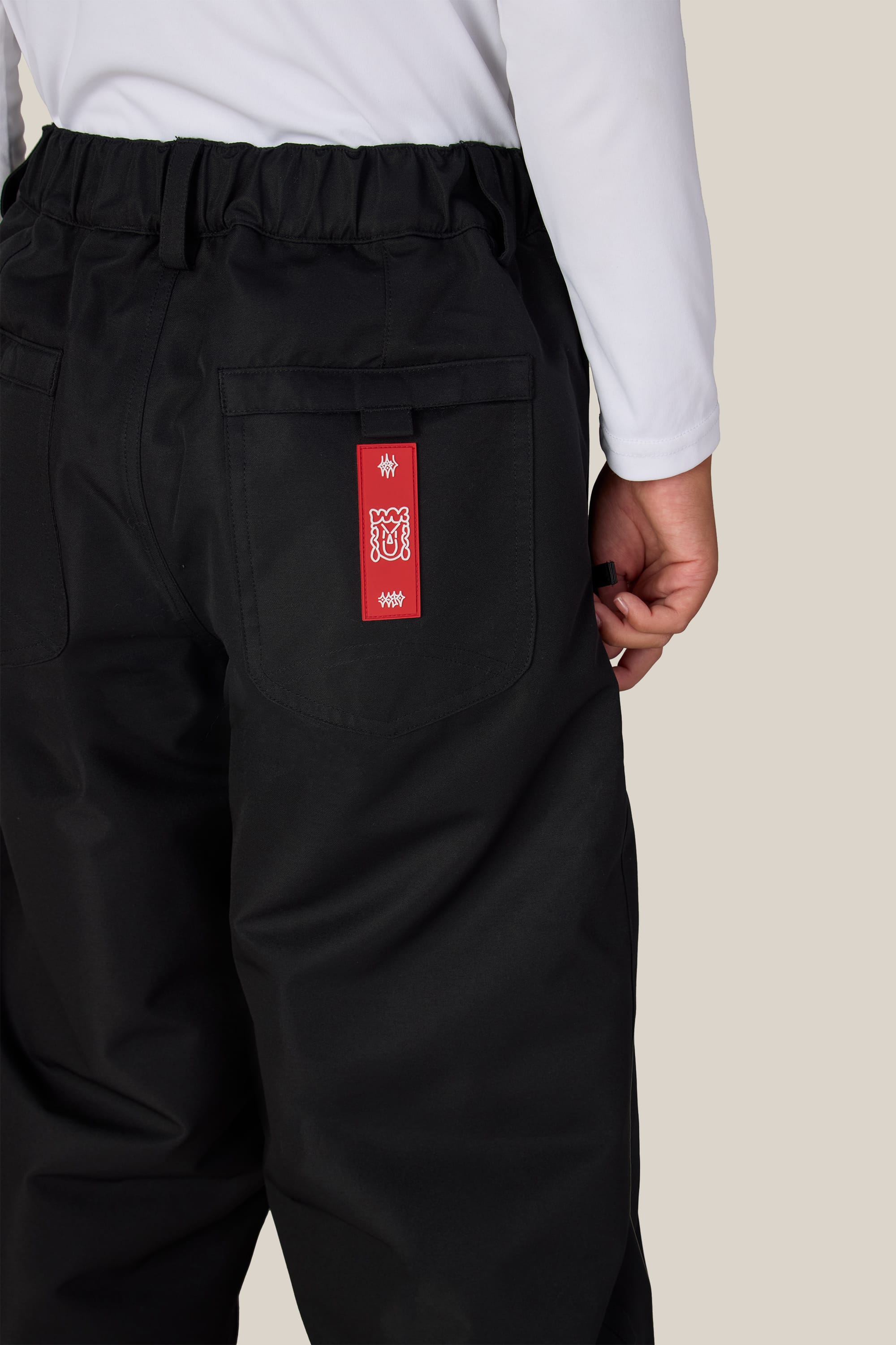 A person wearing 686 Boys’ 686 Dojo® Insulated Pant in black, featuring an elastic waistband, paired with a white long-sleeve shirt; a red tag with white designs is attached to the back right pocket.