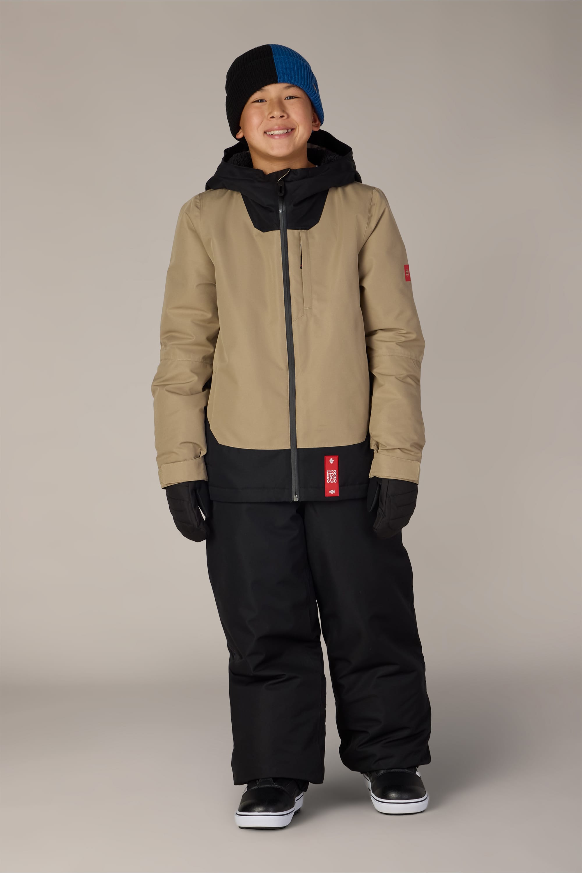 A child wearing a tan and black winter jacket, 686 Boys’ 686 Dojo® Insulated Pant by 686, black gloves, a black and blue beanie, and black shoes stands smiling against a plain light gray background.