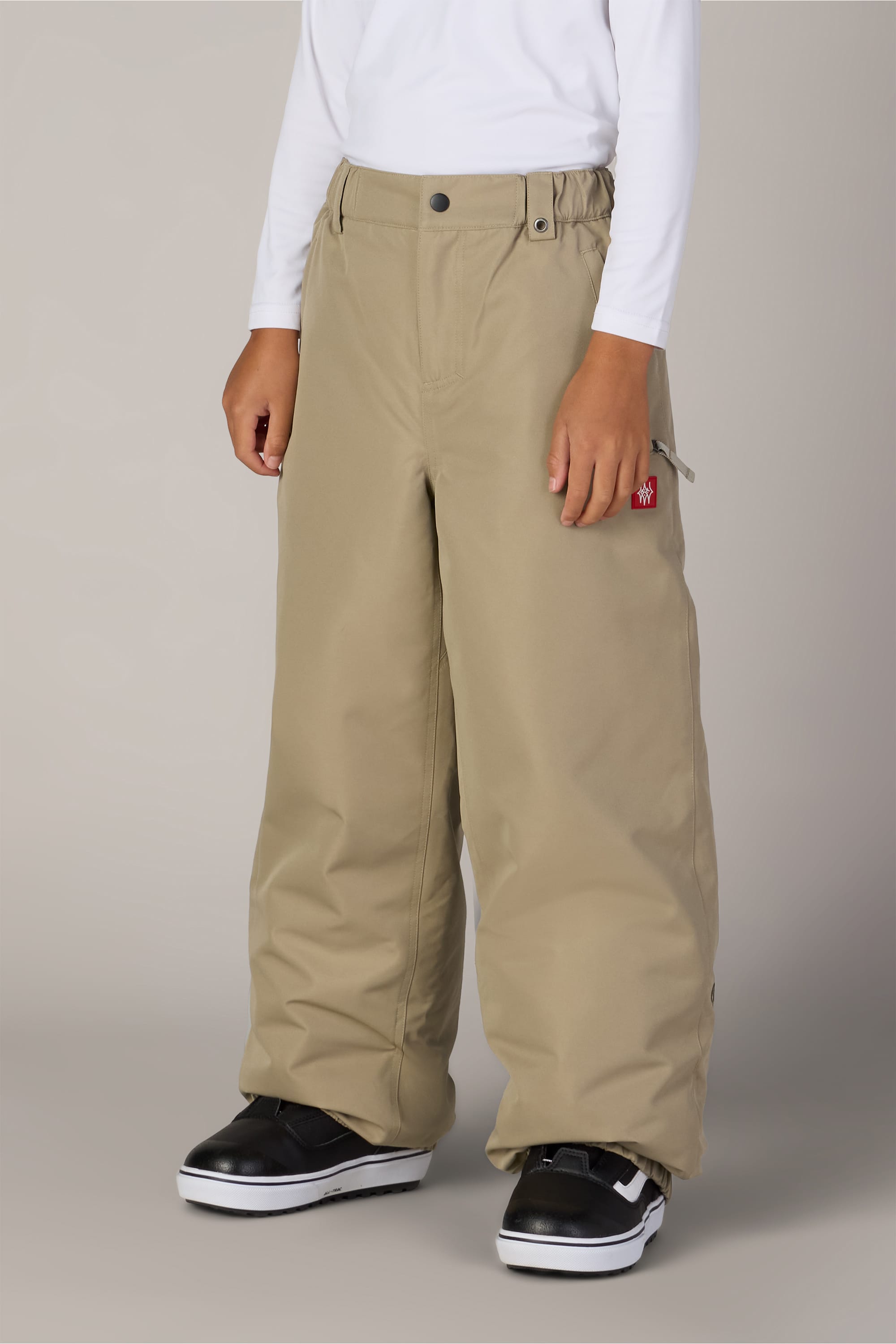 A child is shown standing against a plain light background wearing 686 Boys’ 686 Dojo® Insulated Pant in tan, a white long-sleeve shirt, and black-and-white sneakers. The child’s head is not visible in the image.