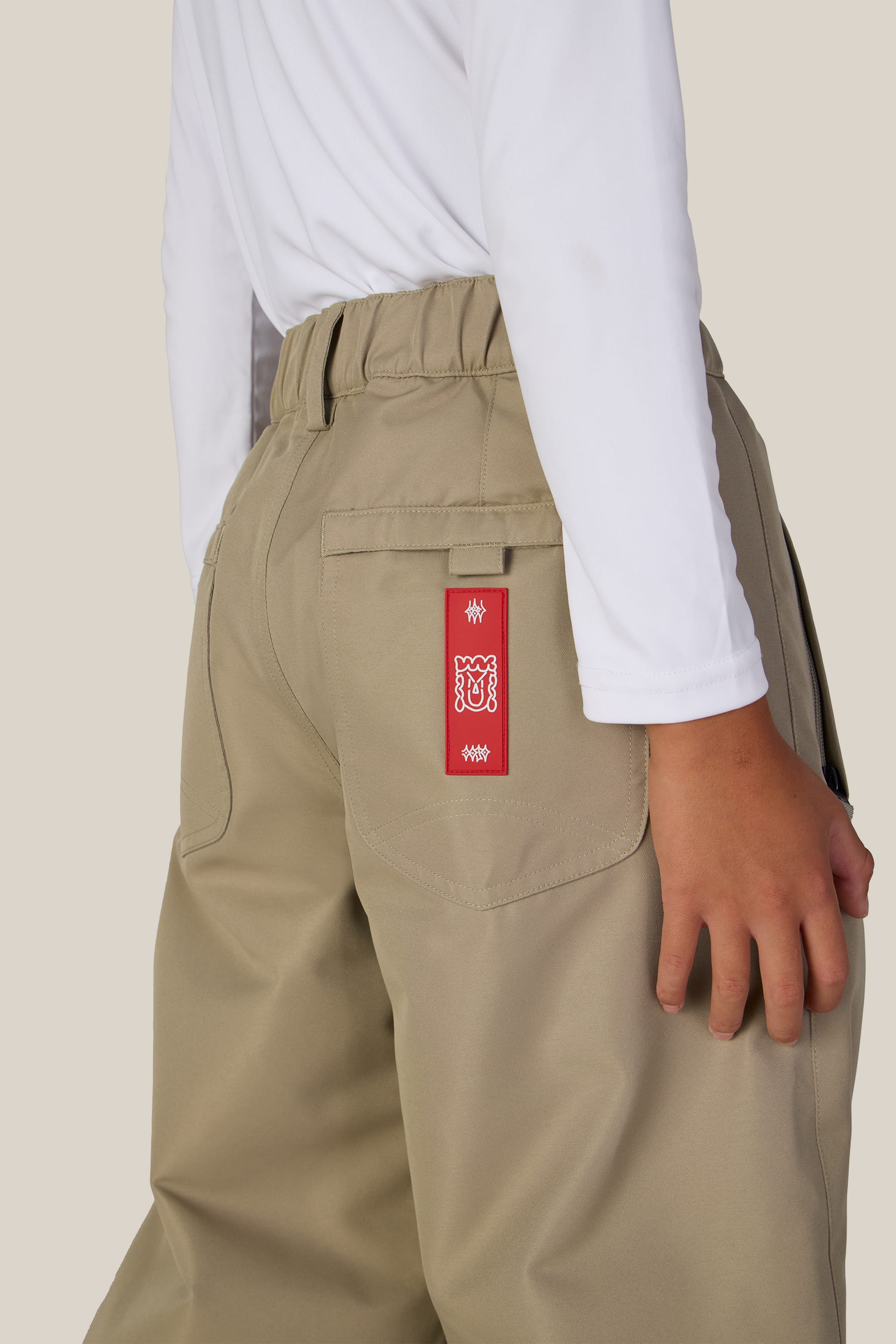 A person wearing the 686 Boys’ 686 Dojo® Insulated Pant in beige, paired with a white long-sleeve shirt, with a red tag featuring white designs hanging from a back pocket.