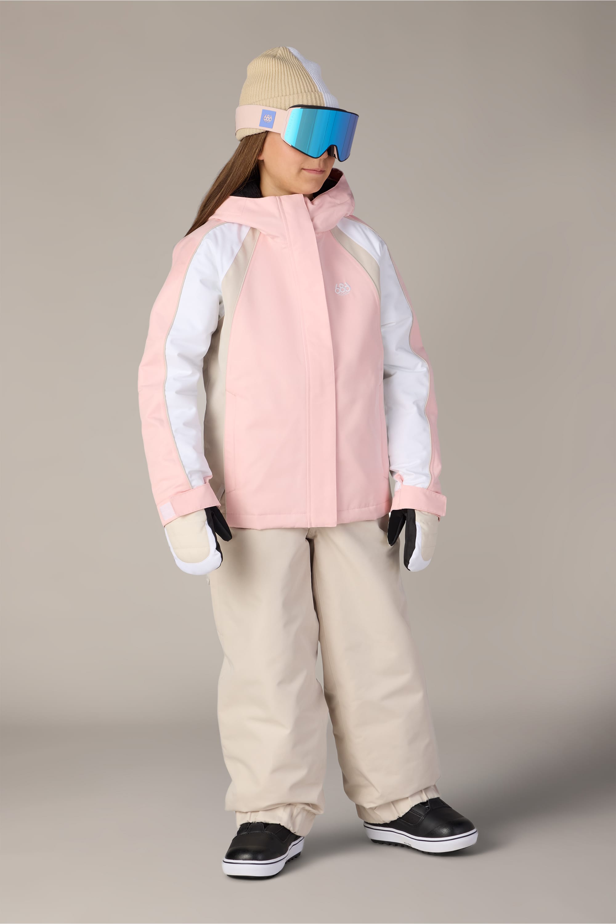 A child is wearing the 686 Girls’ Hologram™ Insulated Jacket in light pink and white by 686, paired with beige snow pants, a beige beanie, blue ski goggles, white gloves, and black snow boots, standing against a plain background.