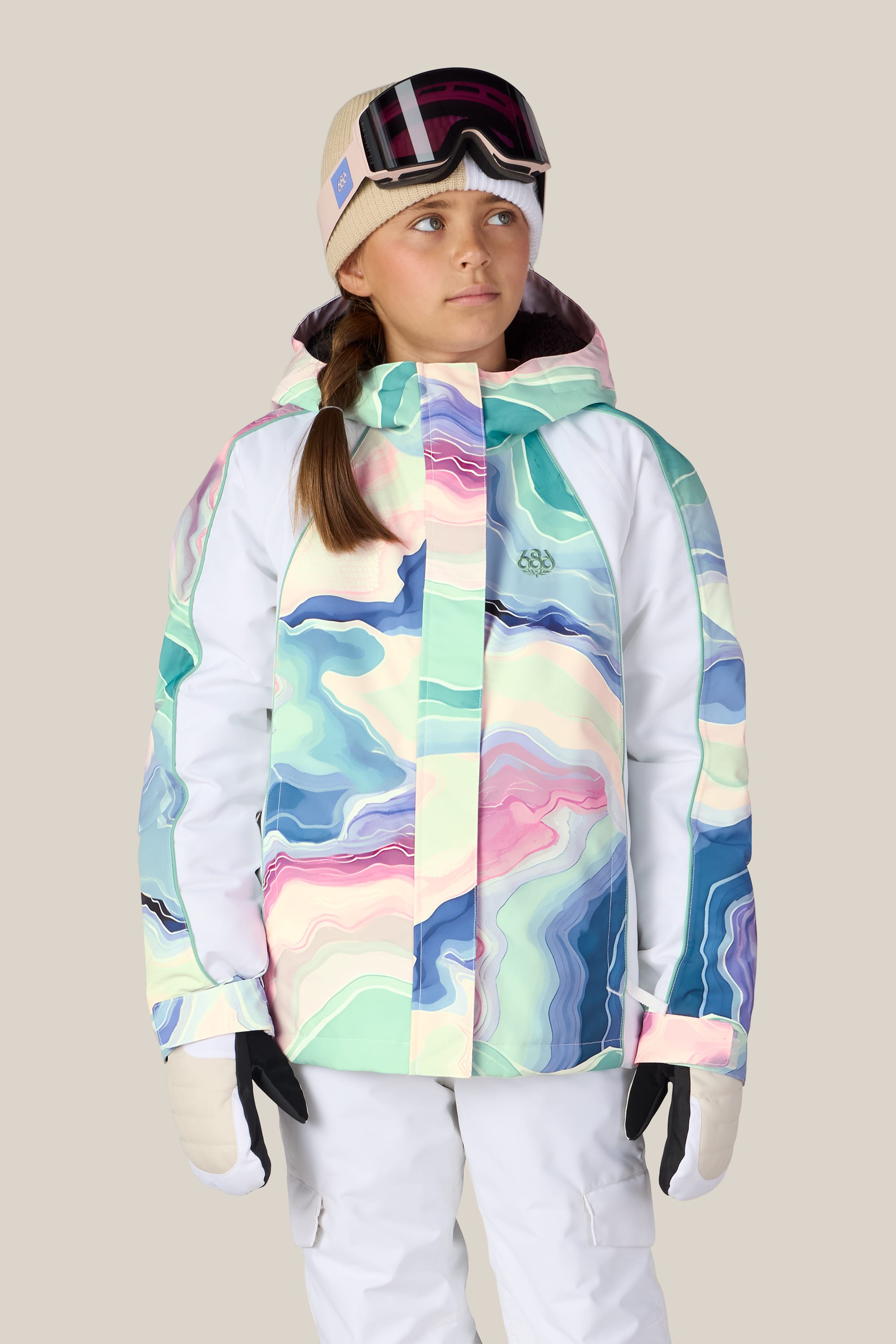 A child wearing the 686 Girls’ Hologram™ Insulated Jacket, featuring a colorful abstract pattern, along with white pants, gloves, a beige beanie, and ski goggles stands against a plain background, looking slightly upward.