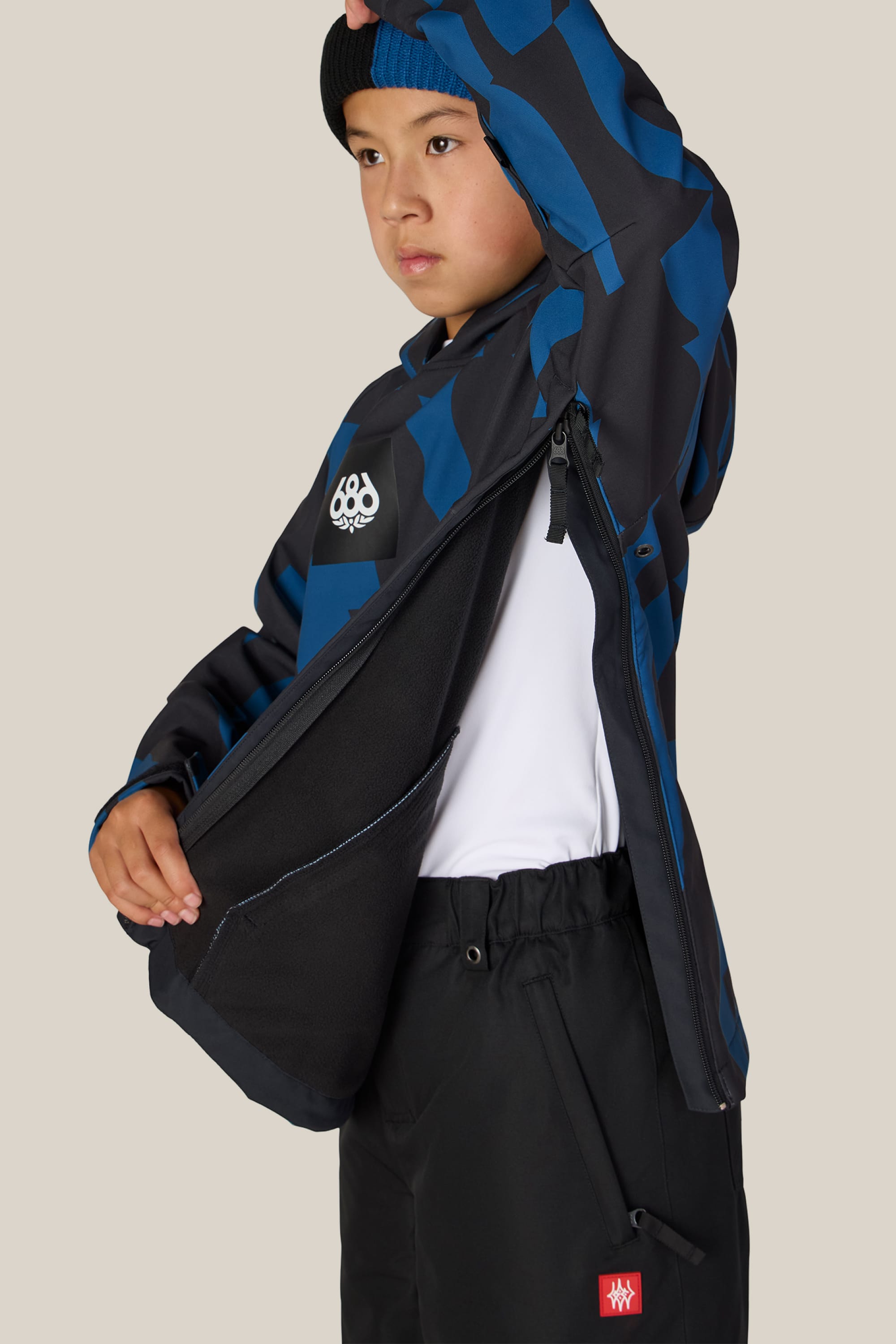A child wearing the 686 Youth Waterproof Hoody by 686, featuring a blue and black pattern, along with black pants and a beanie, unzips the jacket’s side vent to reveal a white shirt underneath. The background is plain beige.