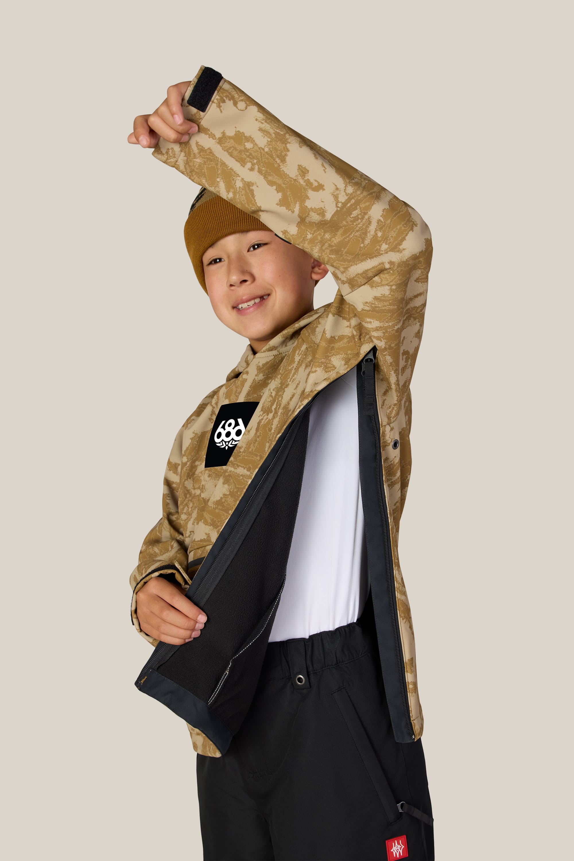 A smiling child wearing the 686 Youth Waterproof Hoody by 686 in tan camo lifts one arm and opens the jacket to reveal its black inner lining, layered over a white shirt and paired with a beanie, against a plain beige background.