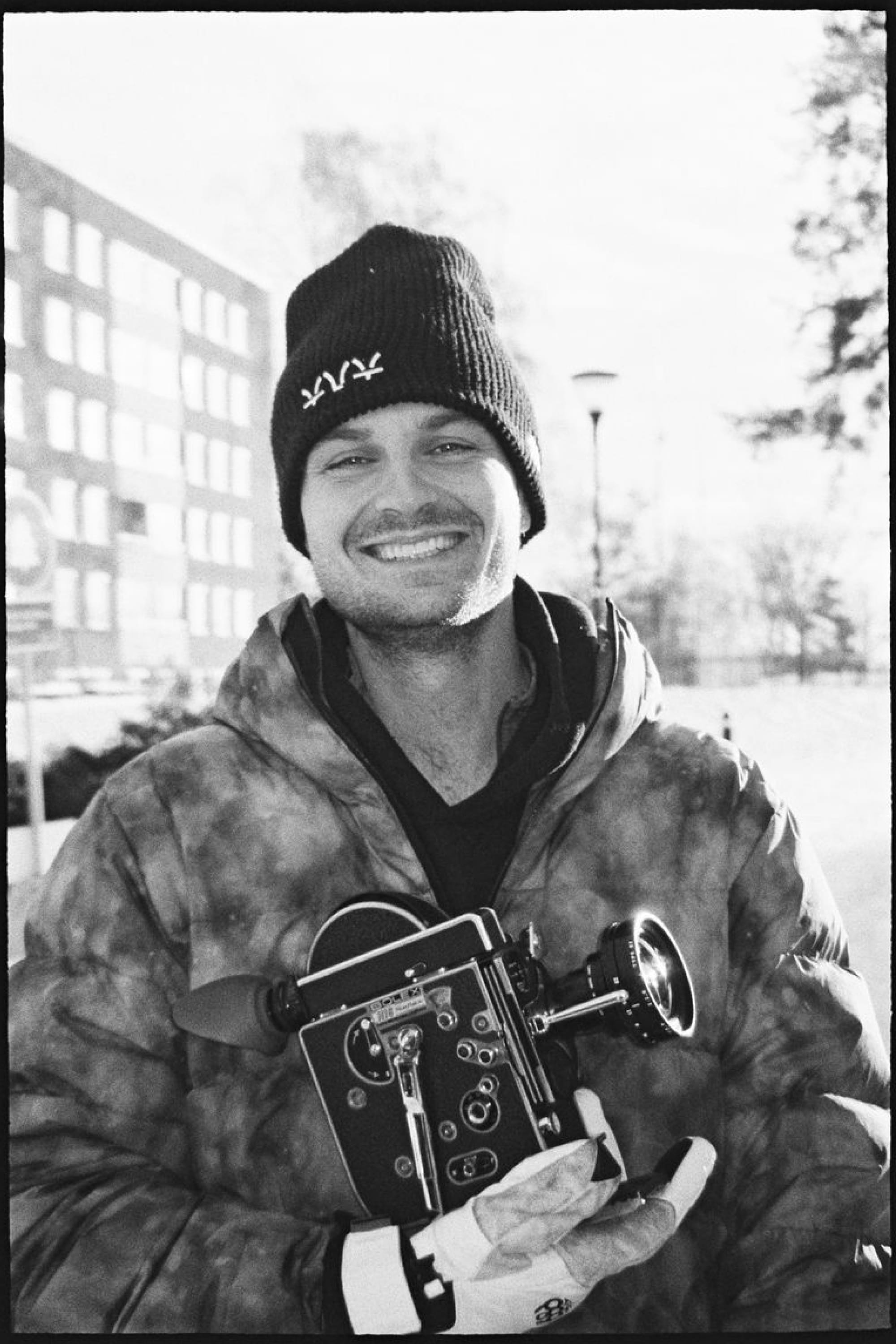 A smiling person wears the 686 Kanji Beanie by 686 and a winter jacket while holding an old-fashioned film camera outdoors on a bright day.
