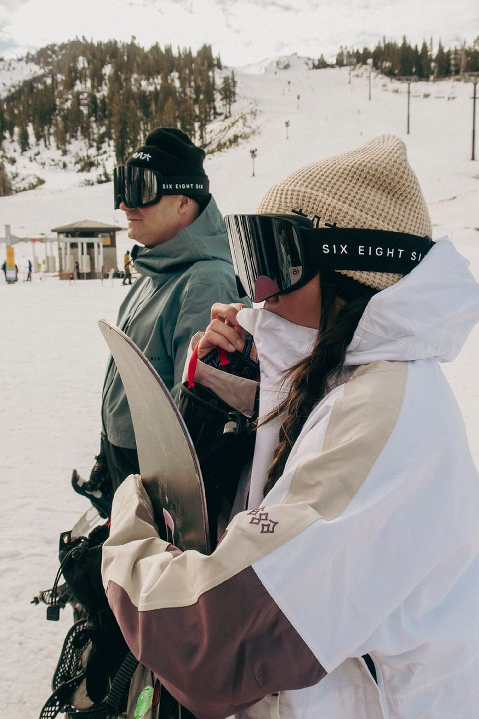 Two people stand in the snow holding snowboards, wearing winter jackets, ski goggles, and 686 Kanji Beanies by 686, with snowy mountains and ski lifts in the background.