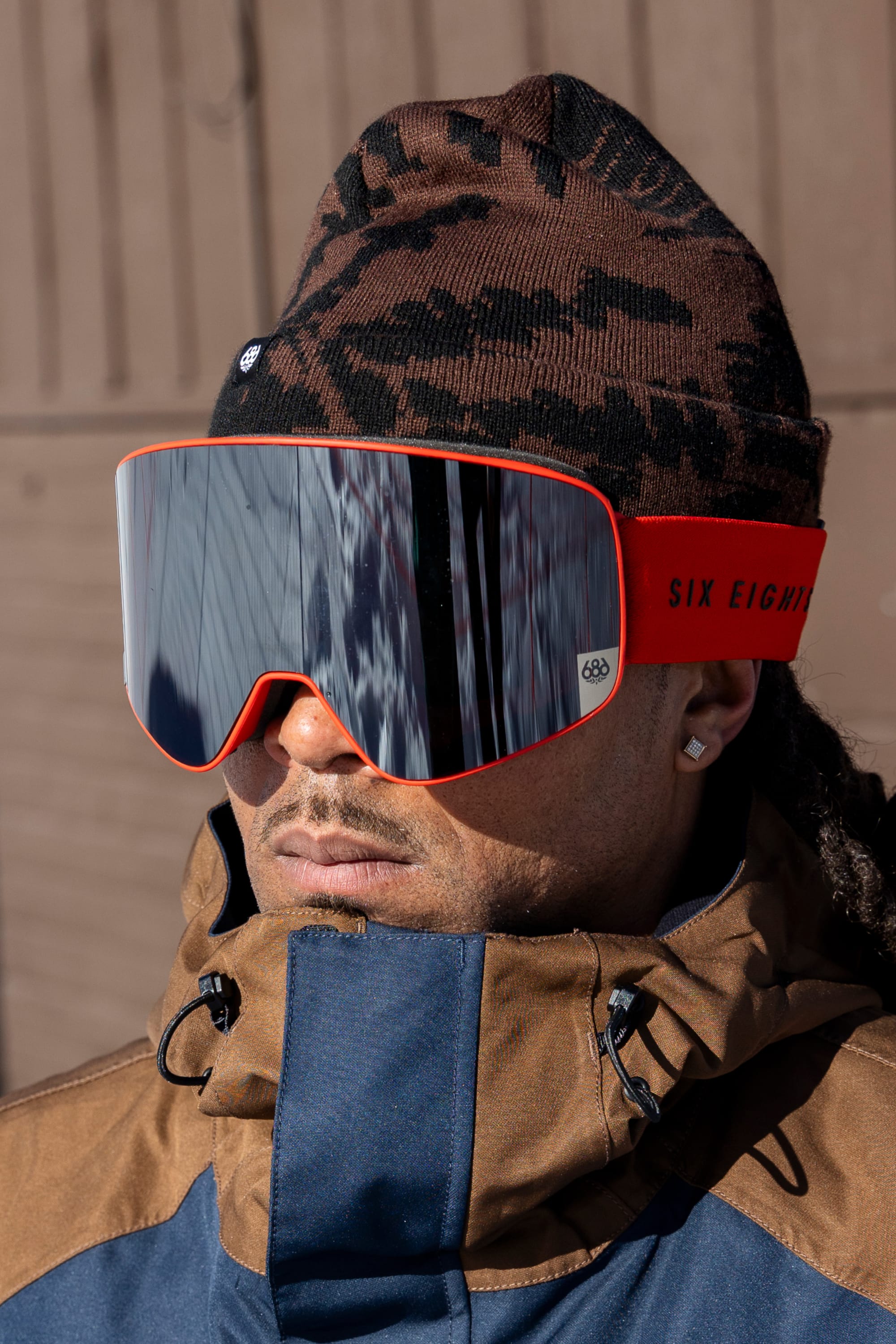 A person wearing the 686 Reverb Beanie by 686, along with large red ski goggles with reflections and a brown and blue jacket, stands outdoors.