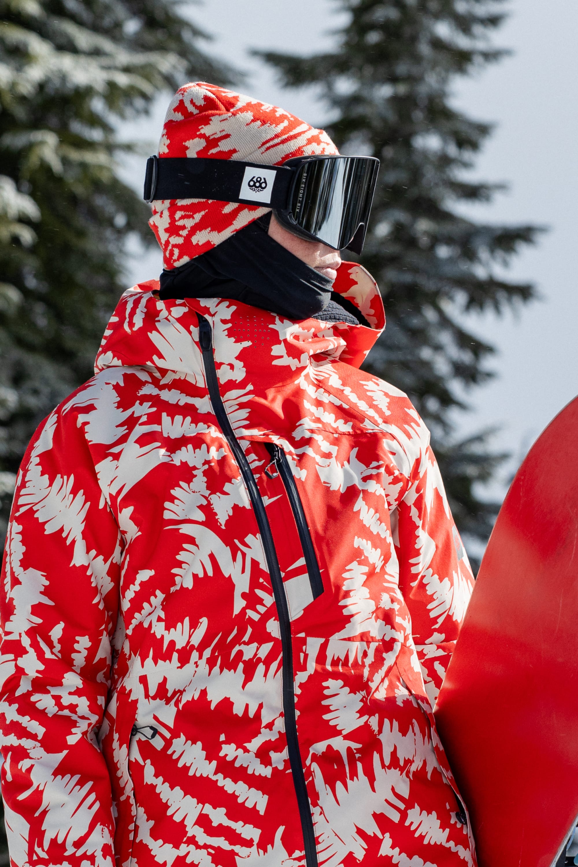 Wearing the 686 Reverb Beanie by 686, a person in a red and white patterned jacket and hat stands outdoors against snow-covered trees. They hold a snowboard, sporting black ski goggles and a face covering.