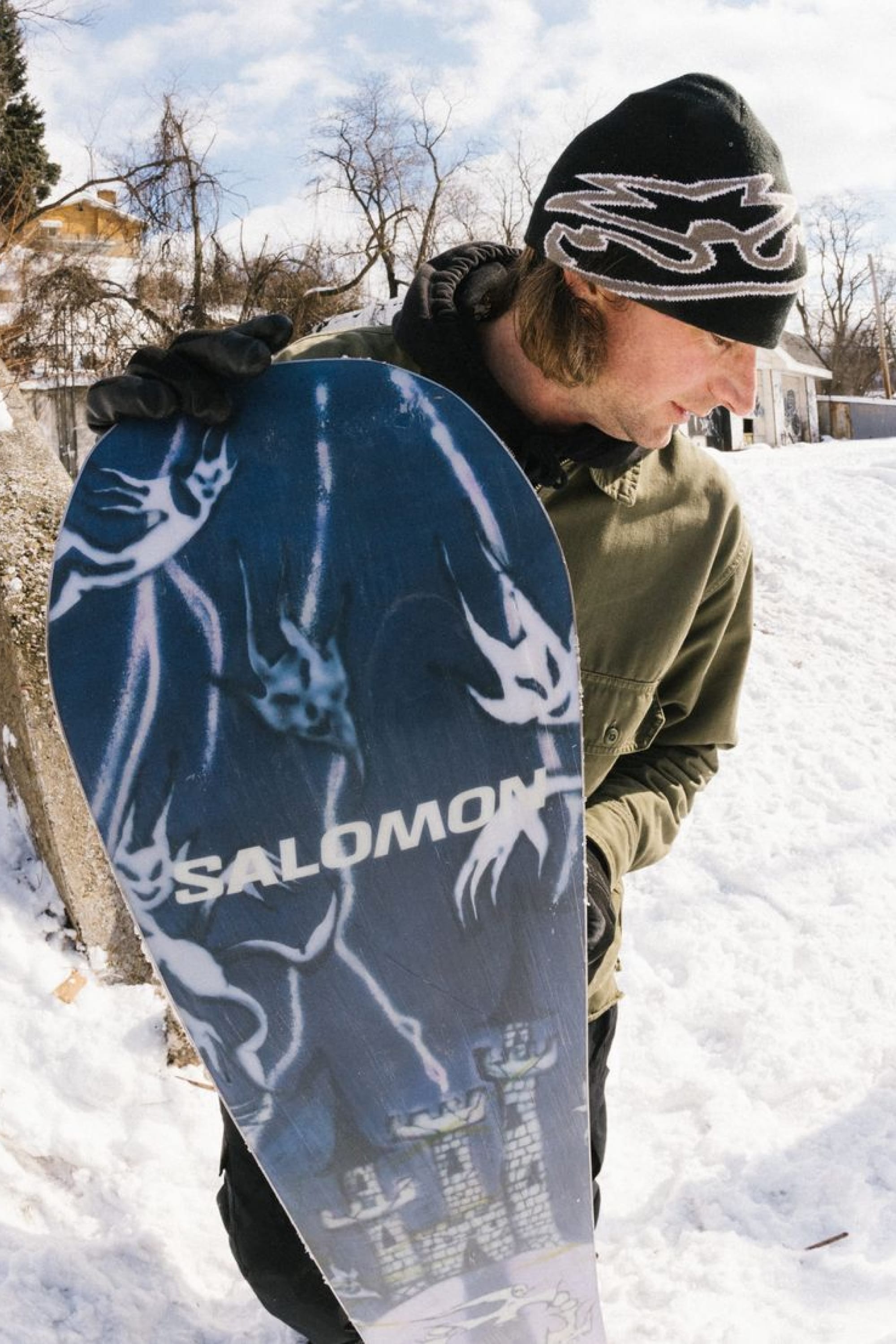Wearing winter clothing and a 686 Jib Skully Beanie by 686, a person holds a Salomon snowboard with a blue and white graphic design while standing on snow near trees and houses.