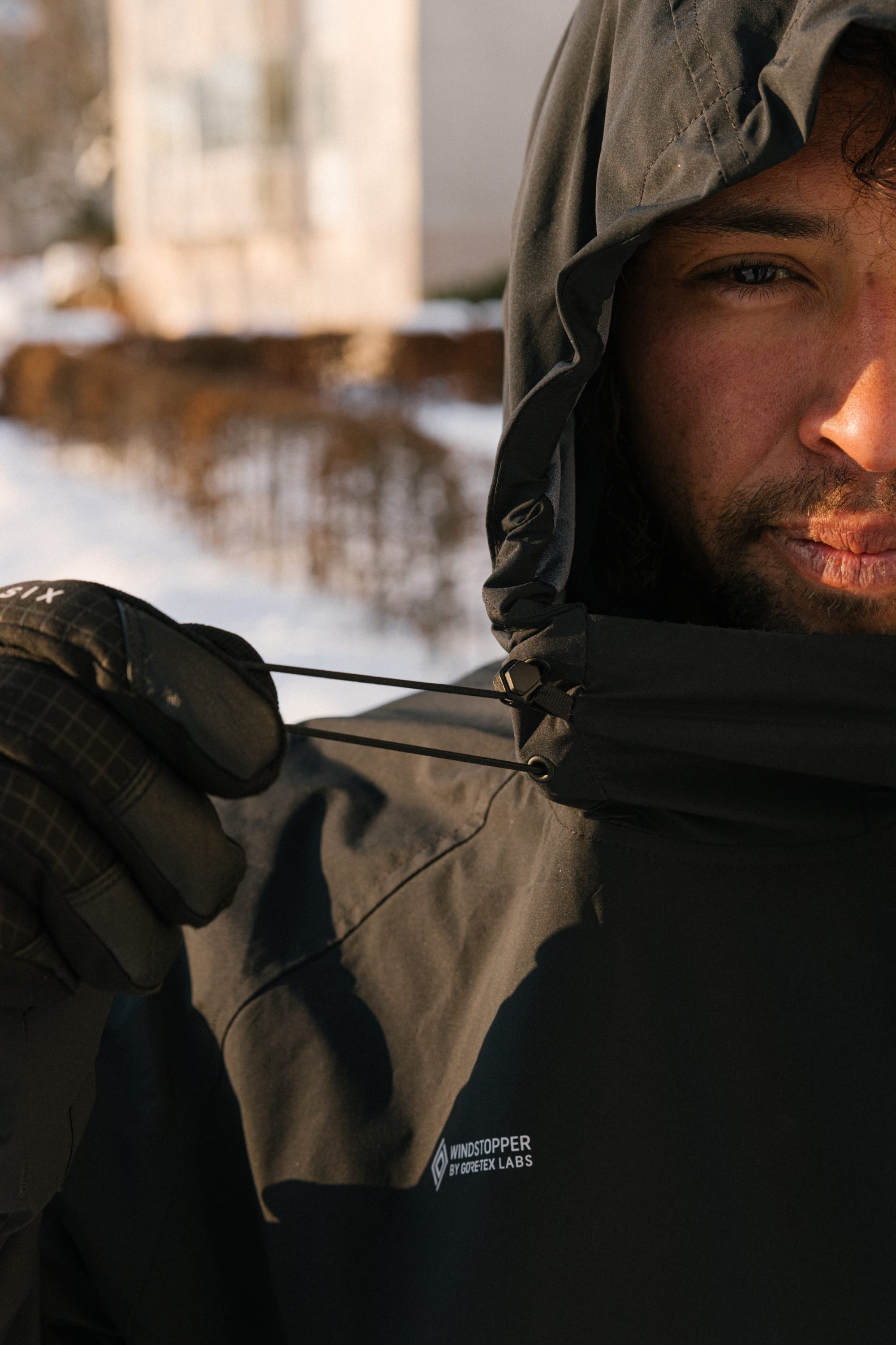 Wearing the 686 Men’s WINDSTOPPER® Pullover Hoody by GORE-TEX® LABS, a person pulls the drawstring tight on their dark hooded jacket, partially obscuring their face. Behind them, a snowy outdoor scene and blurred buildings complete the background.