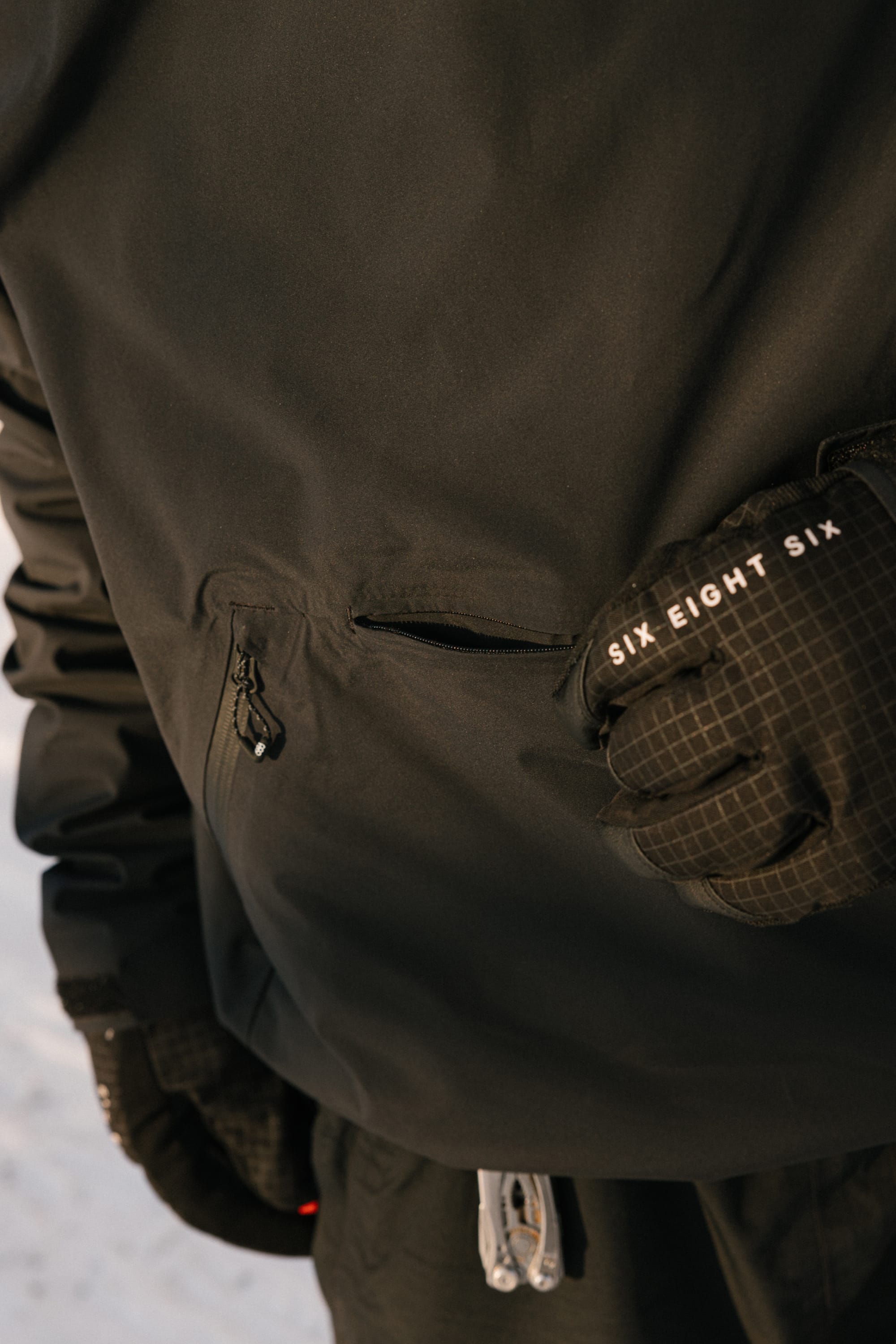 A person wearing the 686 Men’s WINDSTOPPER® Pullover Hoody by GORE-TEX® LABS and black gloves, standing outdoors in the snow. One glove features the text SIX EIGHT SIX, and a zippered pocket on the hoody is visible.