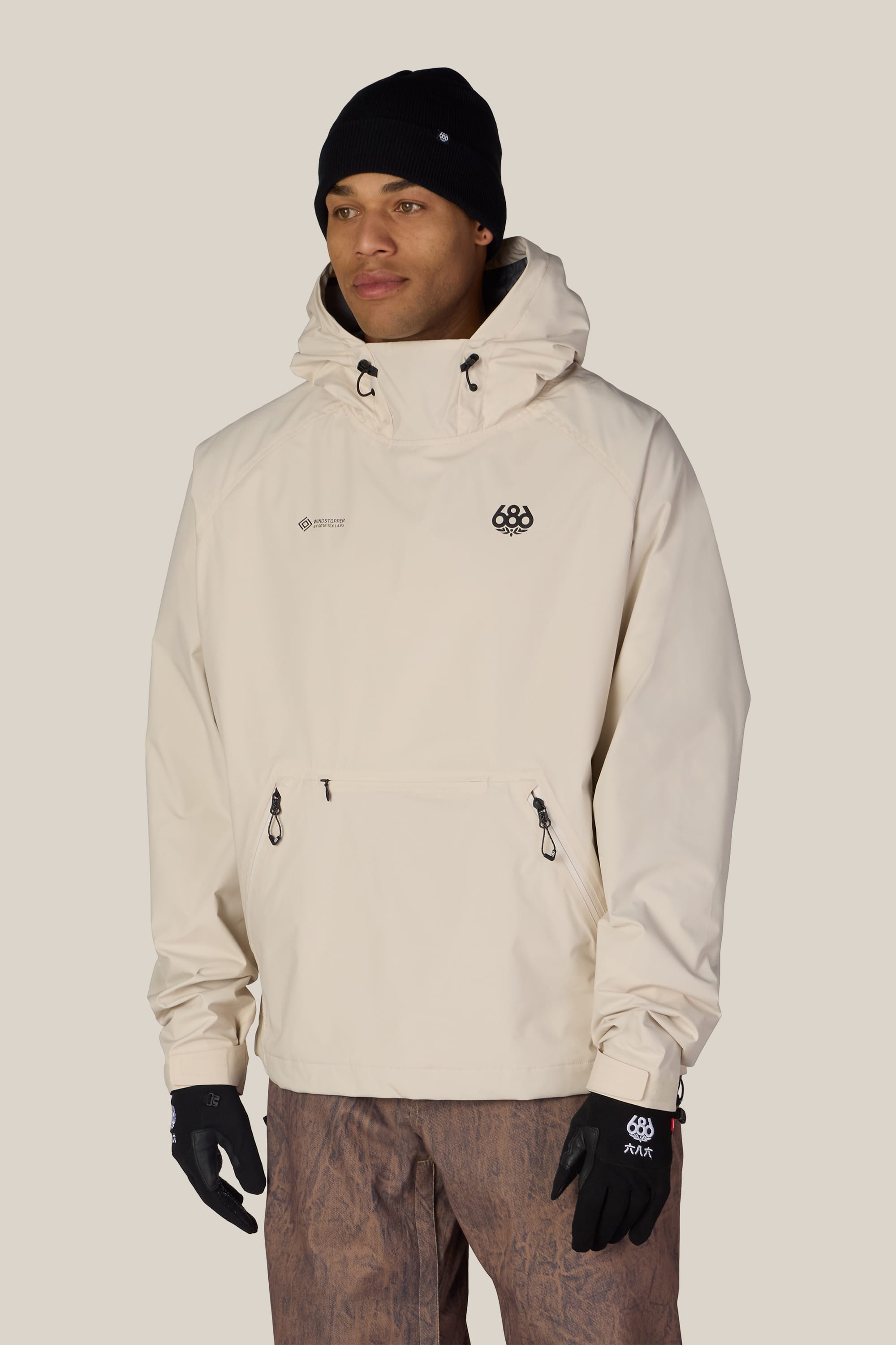A person wearing the 686 Men’s WINDSTOPPER® Pullover Hoody by GORE-TEX® LABS in cream, along with a black beanie and black gloves, stands against a plain background. The hoody features a front zipper pocket and small 686 logos on the chest and gloves.