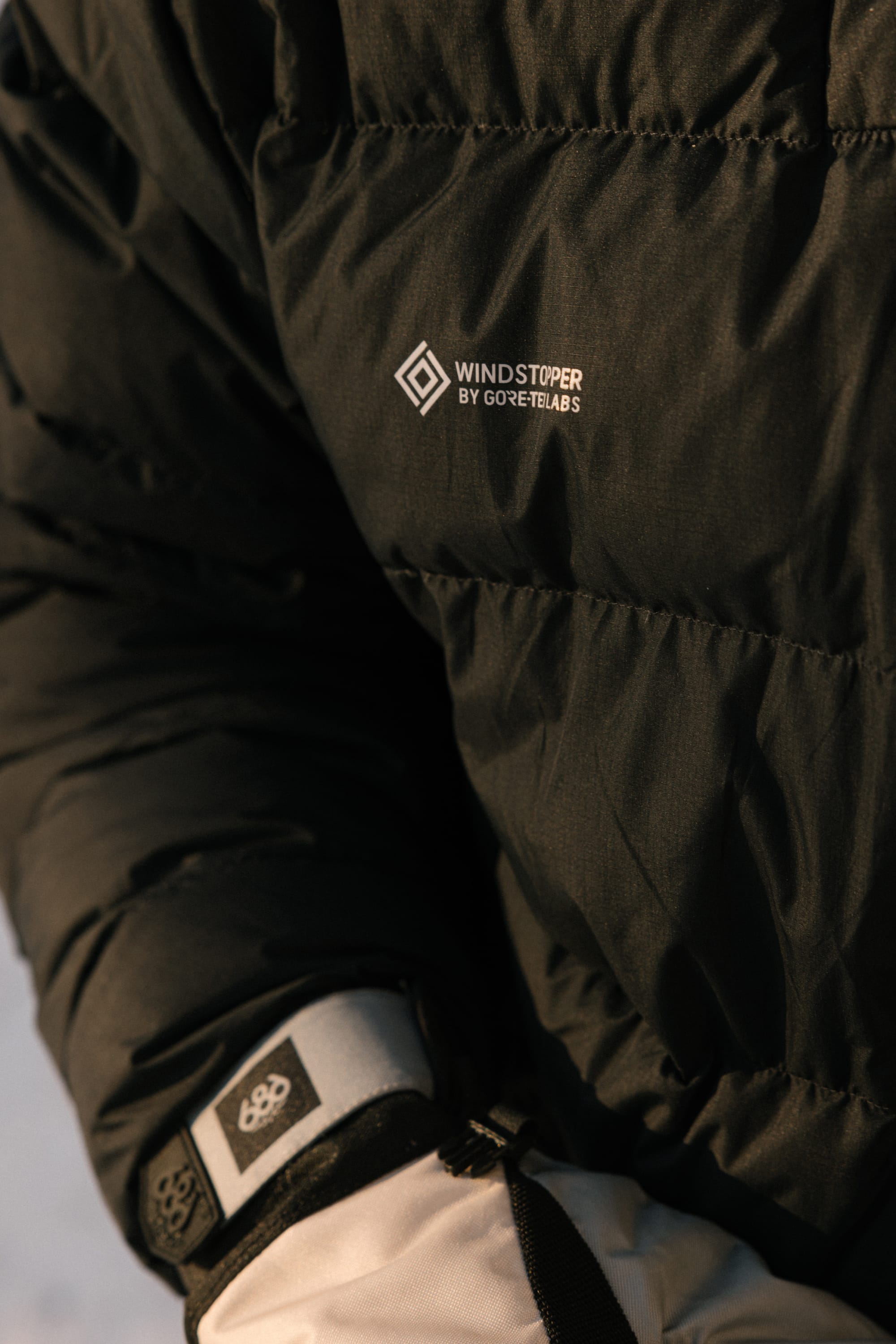 Close-up of the 686 Men’s WINDSTOPPER® Pyro™ Down Puffer Jacket by GORE-TEX® LABS in black, showing insulation details and the Windstopper logo, alongside part of a white glove featuring the 686 logo on the wrist.