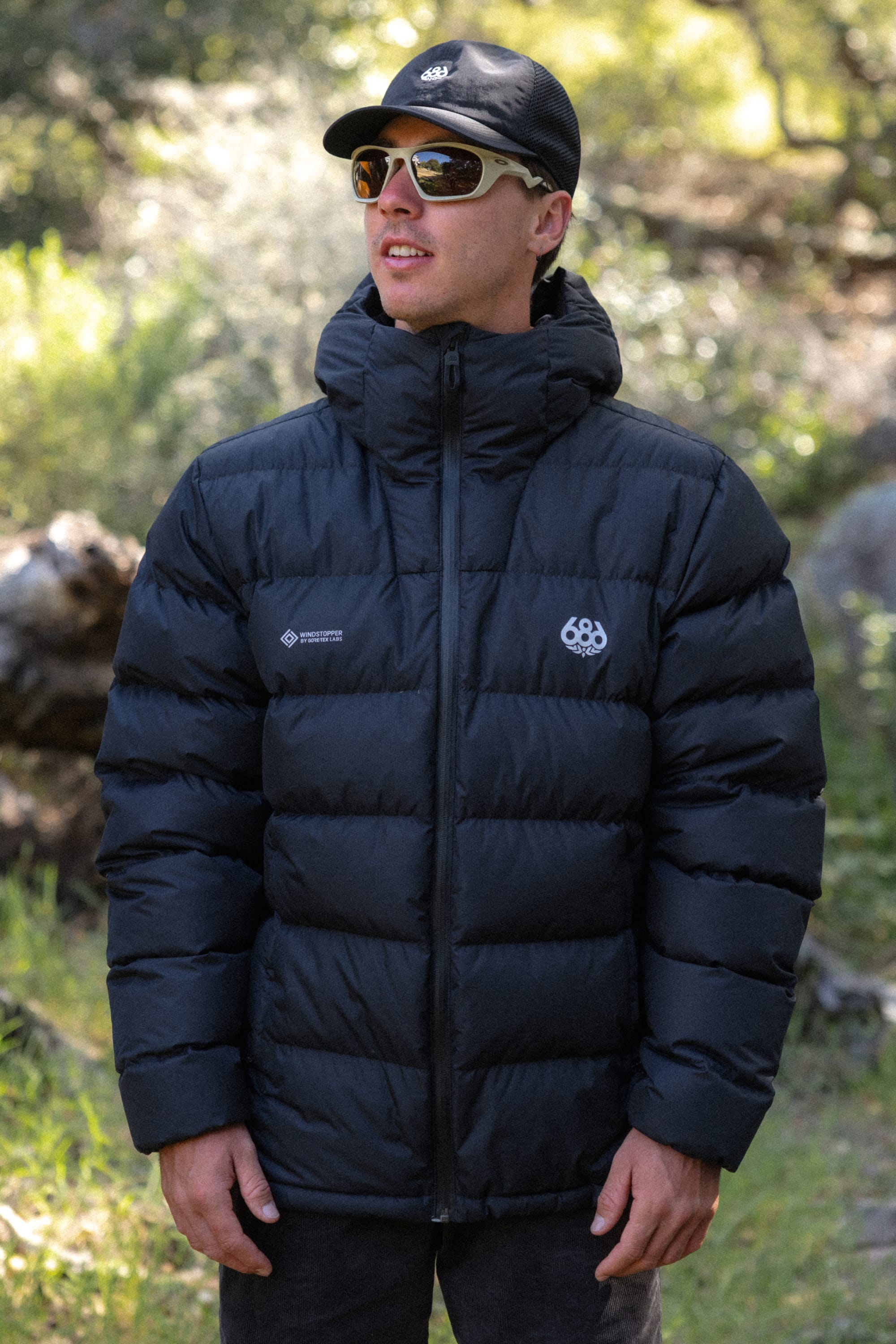 Wearing the 686 Men’s WINDSTOPPER® Pyro™ Down Puffer Jacket by GORE-TEX® LABS, a person stands outdoors in a sunlit, wooded area, looking slightly upward and smiling while also sporting a black cap and sunglasses.
