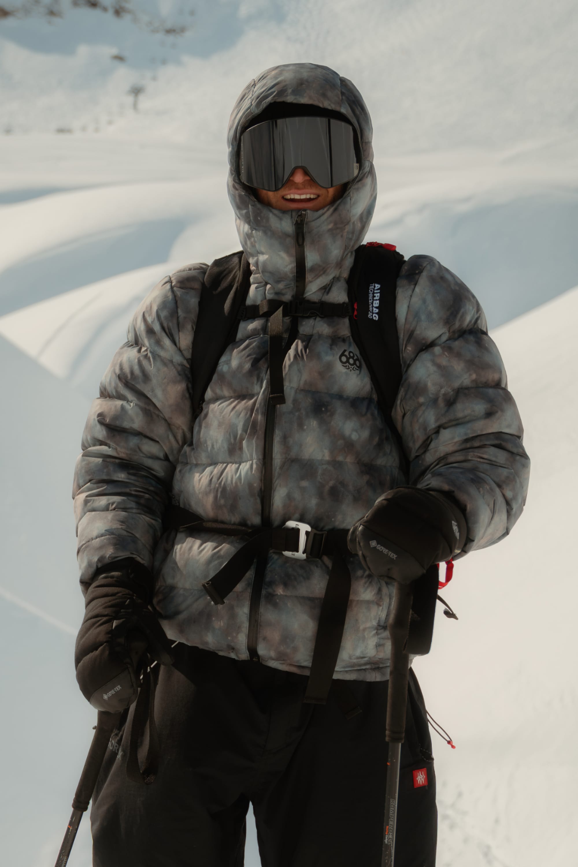 Wearing the 686 Men’s WINDSTOPPER® Pyro™ Down Puffer Jacket by GORE-TEX® LABS, a person stands outdoors in a snowy landscape with black gloves, ski goggles, and a backpack, holding ski poles and smiling.