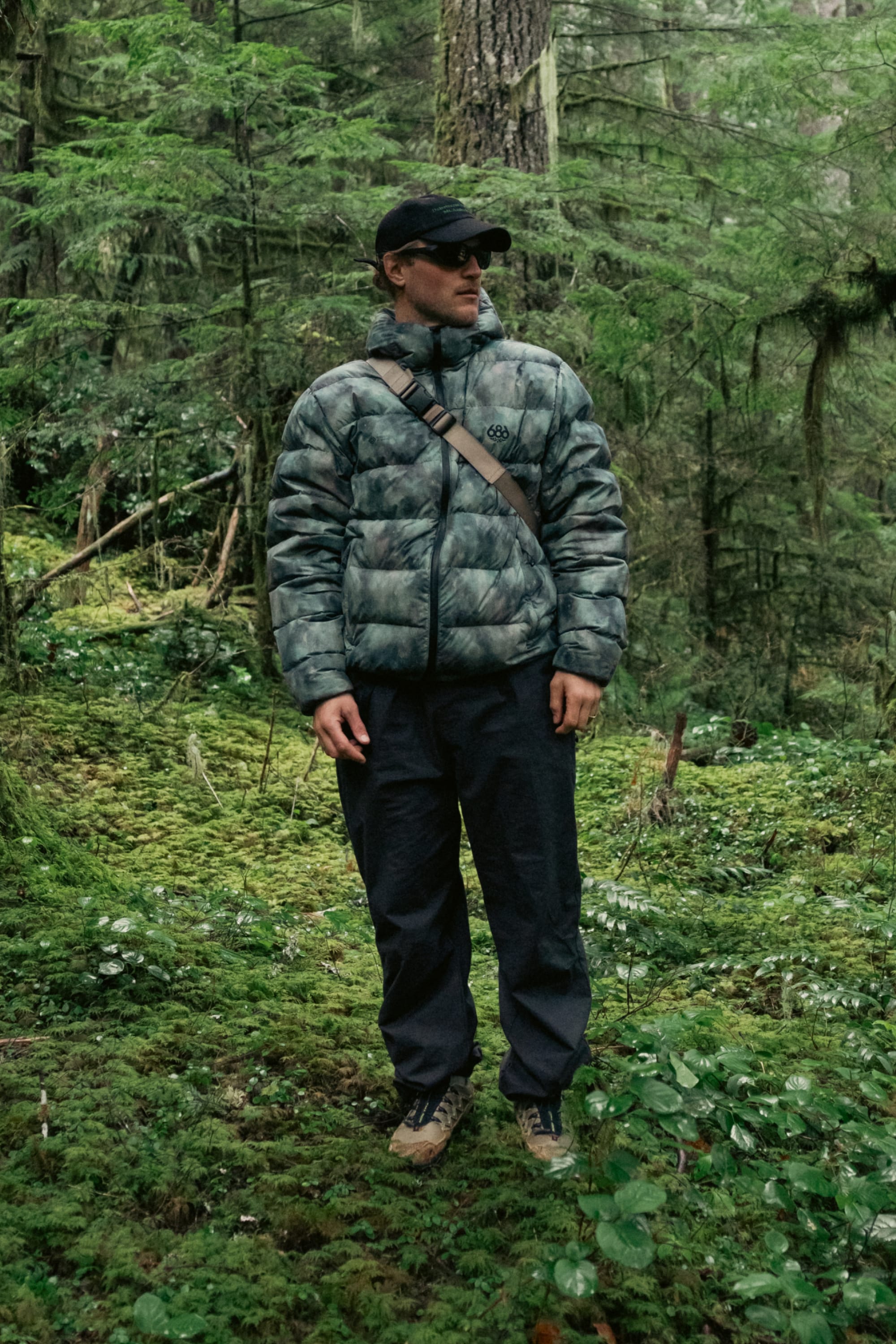 A person wearing a cap, sunglasses, dark pants, a crossbody bag, and the 686 Men’s WINDSTOPPER® Pyro™ Down Puffer Jacket by GORE-TEX® LABS stands in a lush, green forest surrounded by trees and moss.