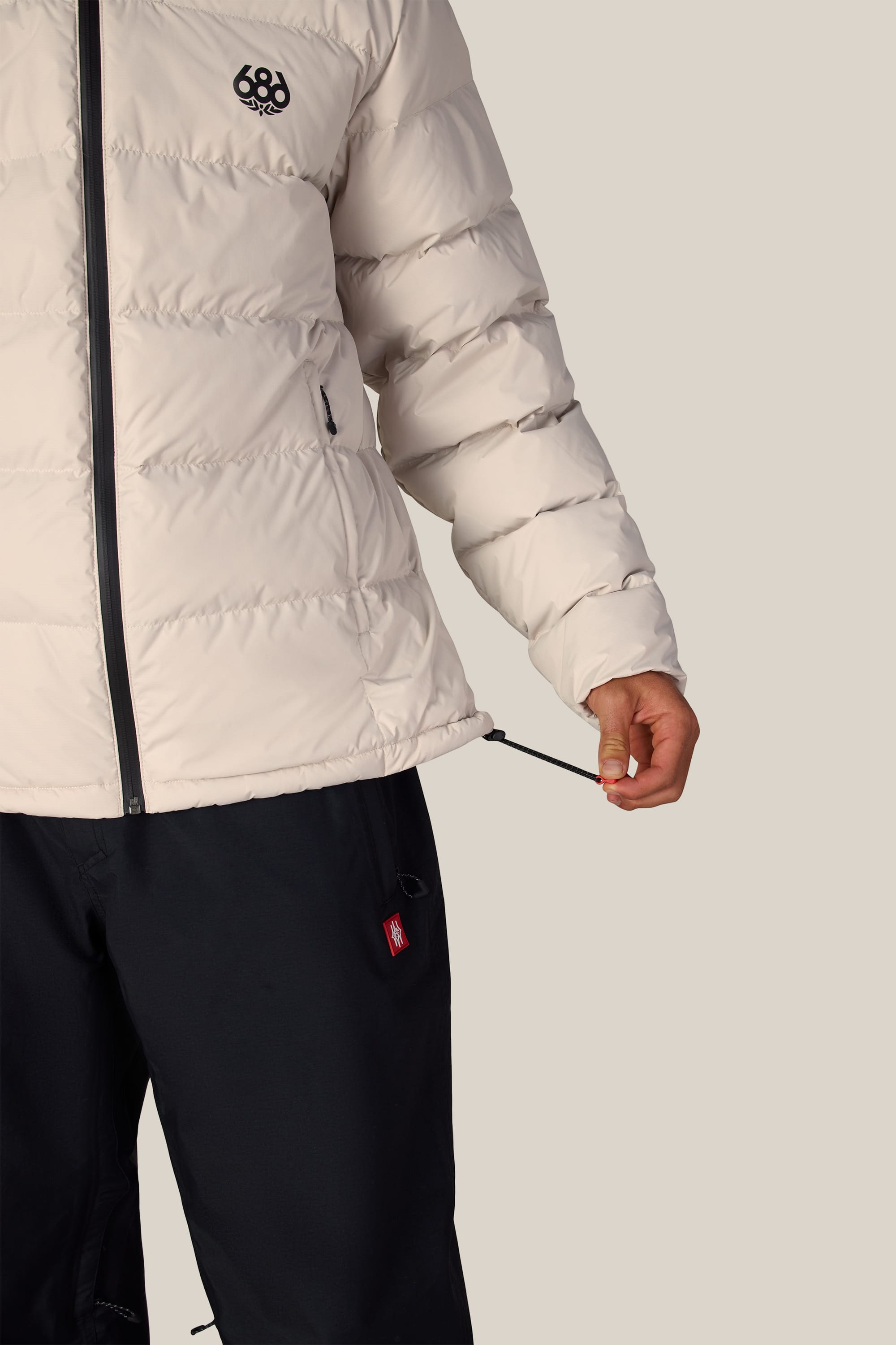 A person wearing the 686 Men’s WINDSTOPPER® Pyro™ Down Puffer Jacket by GORE-TEX® LABS in a light color and black pants adjusts the jacket’s drawstring at the hem, with only their torso and one arm visible against a plain background.