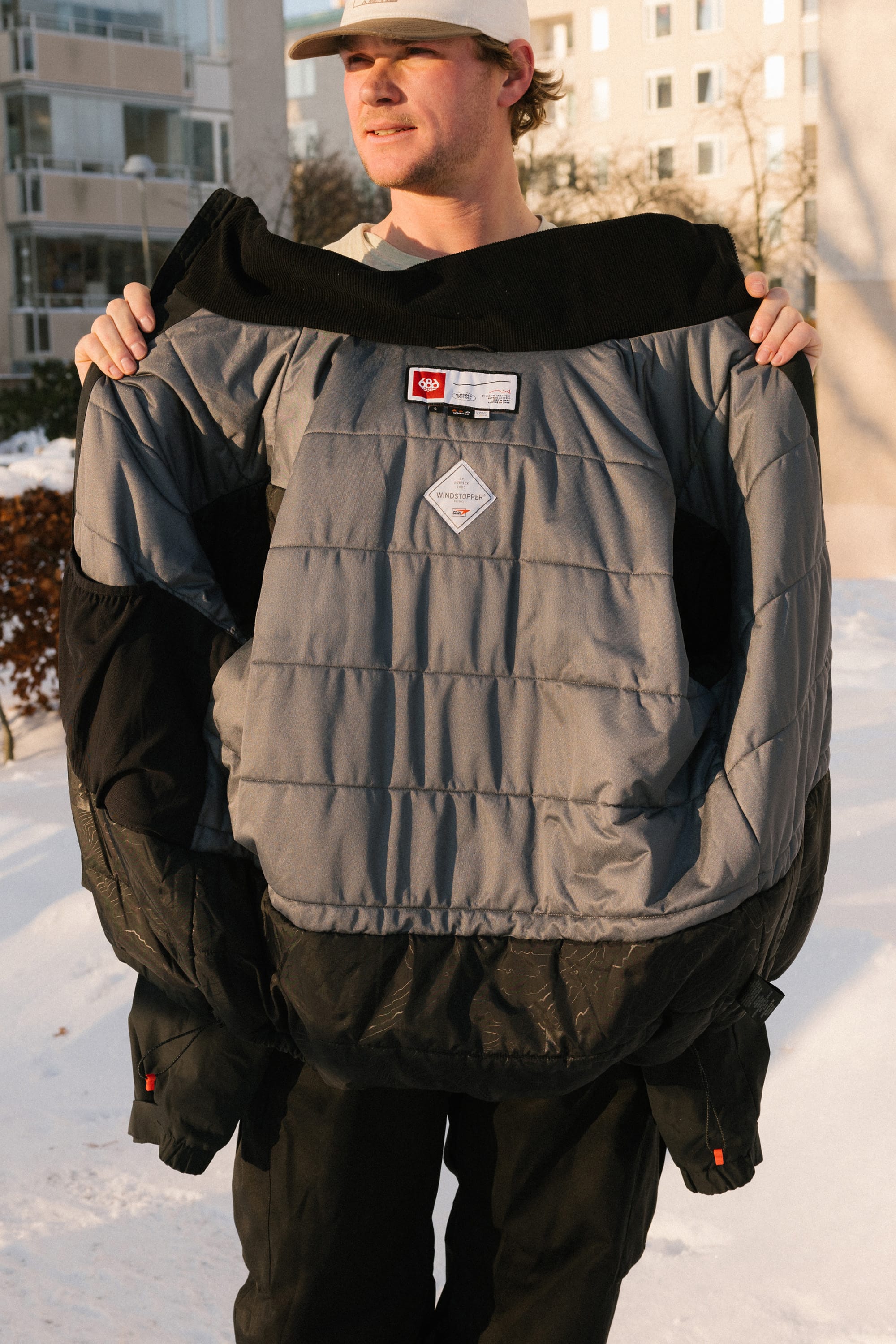A person outdoors in winter holds open a large gray and black 686 Men’s WINDSTOPPER® Guideline Insulated Jacket by GORE-TEX® LABS, revealing its insulated interior. Snow and apartment buildings can be seen in the background.