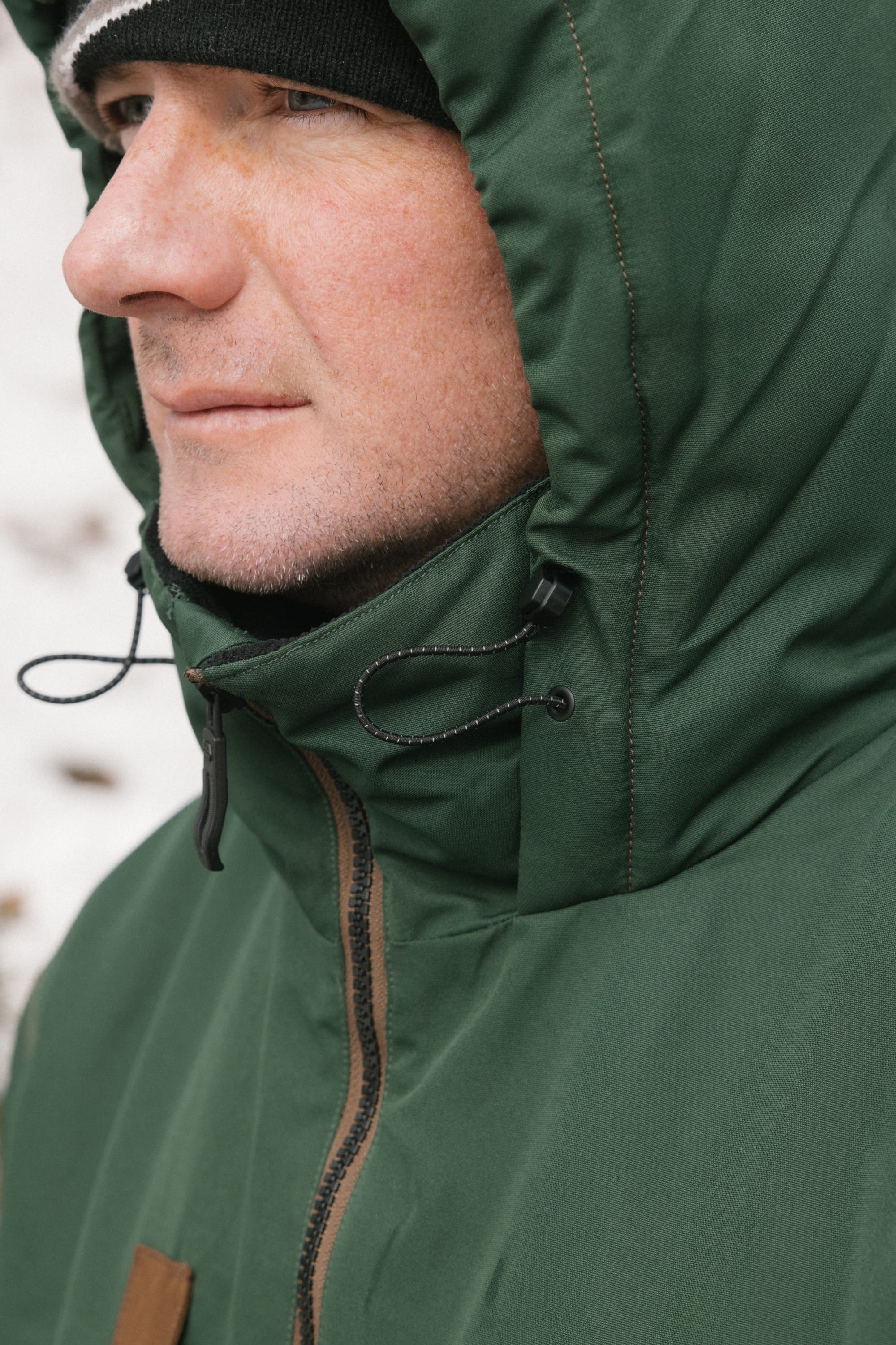 A person wearing the 686 Men’s WINDSTOPPER® Guideline Insulated Jacket by GORE-TEX® LABS stands outside, facing left. They are also wearing a black-and-white beanie, with their face partially visible and snow blurred in the background.