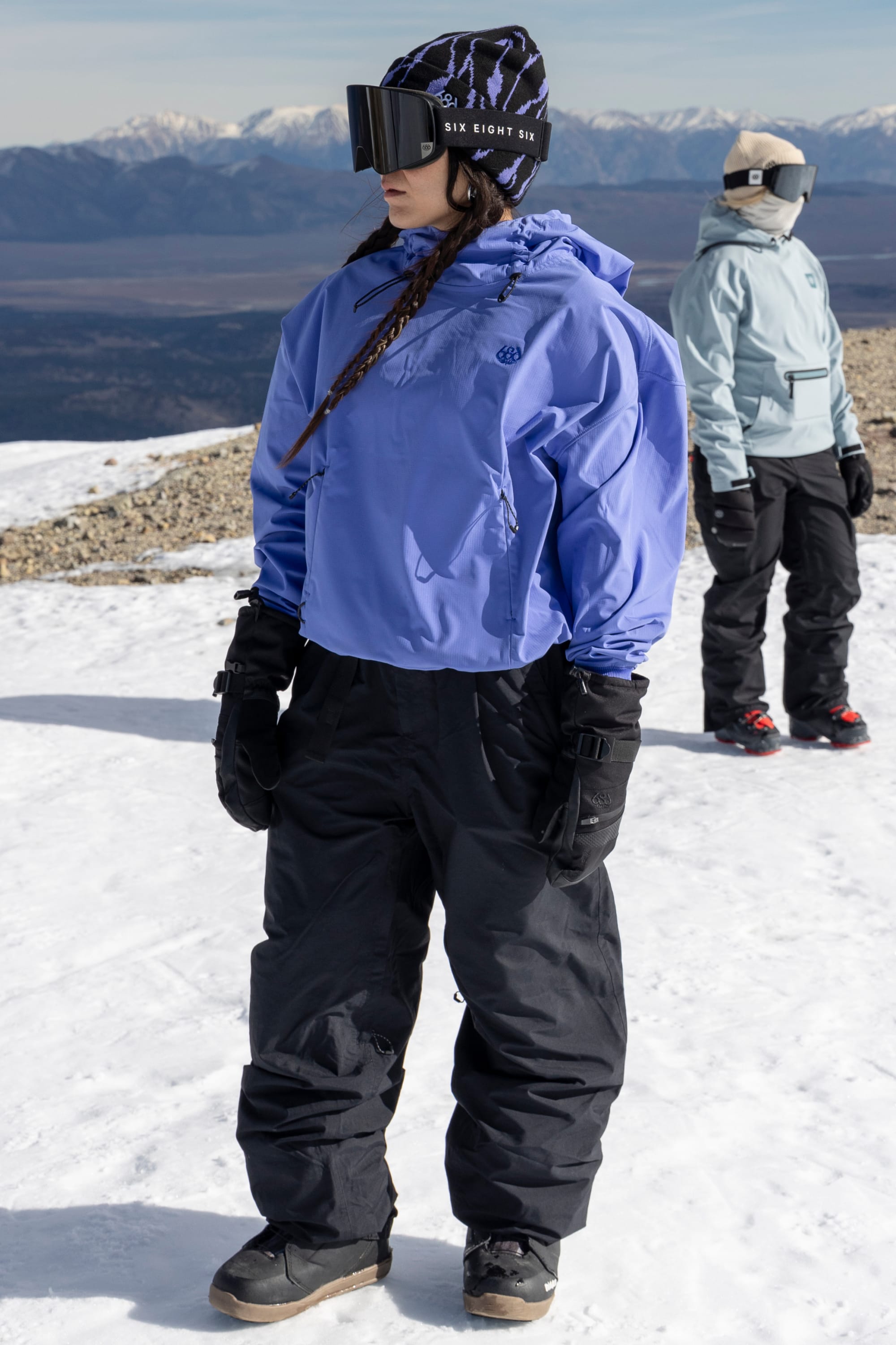 Two people stand on snowy ground in winter gear. The person in front wears the 686 Women’s Waterproof Team Crop Hoody in purple, black pants, gloves, and ski goggles. Mountains are visible in the background under a clear sky.