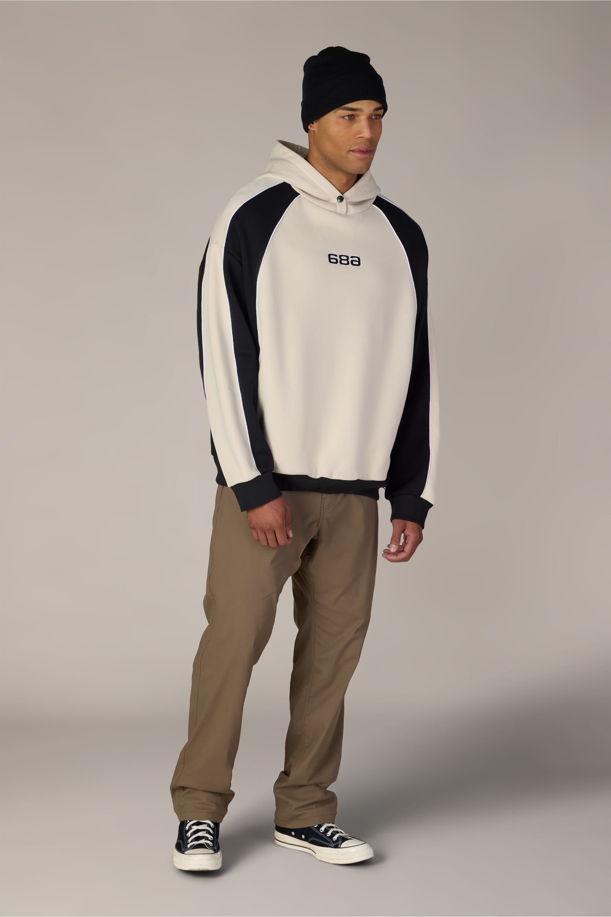 A person stands against a plain background wearing a black beanie, khaki pants, and black sneakers, paired with the 686 Legacy Premium Heavyweight Hoody by 686—a white and black men’s sweatshirt featuring “688” on the chest and crafted from a soft cotton sweatshirt blend.