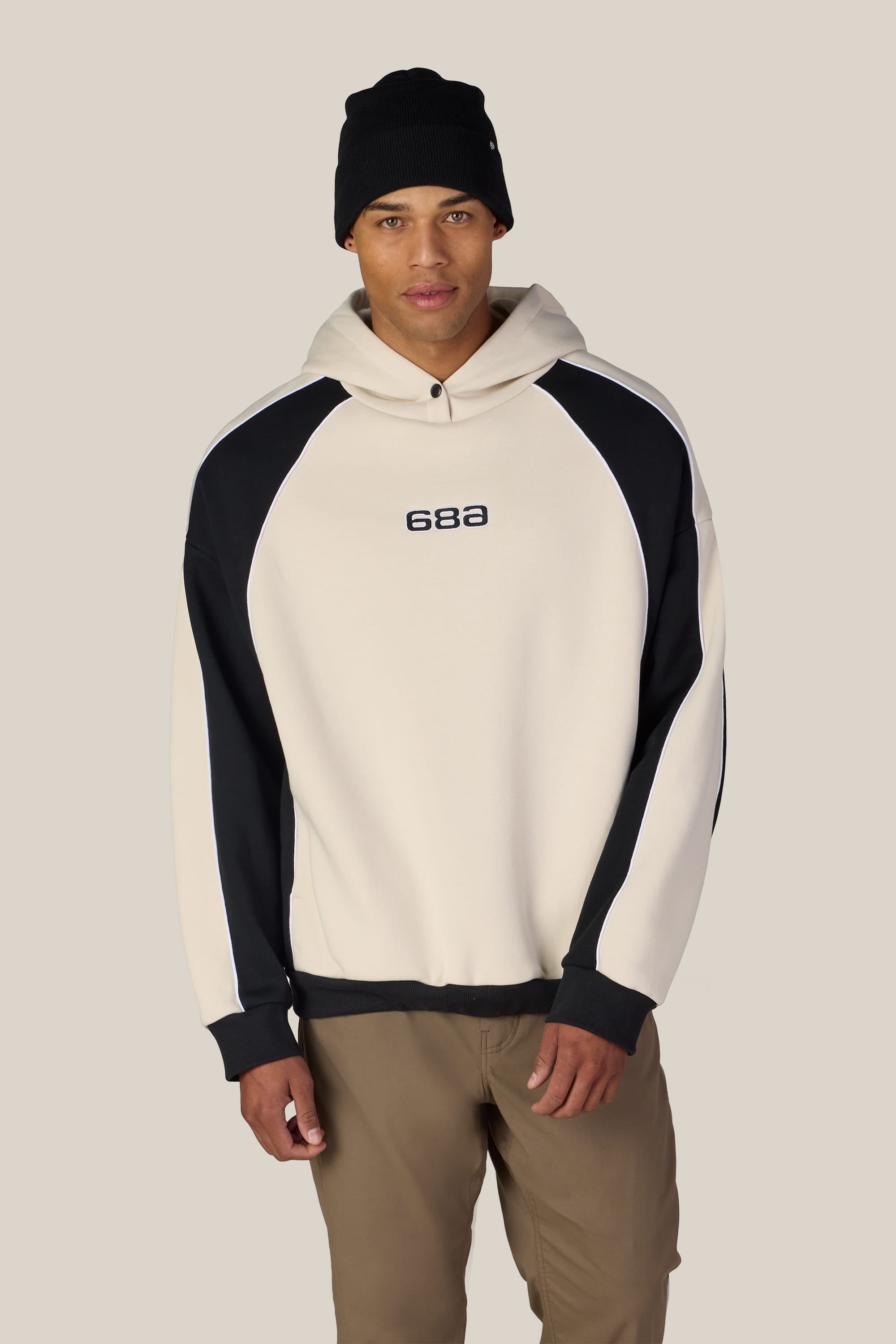 A person wearing a black beanie, brown pants, and the 686 Legacy Premium Heavyweight Hoody by 686—a beige and black men’s sweatshirt with “686” text on the chest, crafted from soft cotton sweatshirt fabric—stands against a plain light background.
