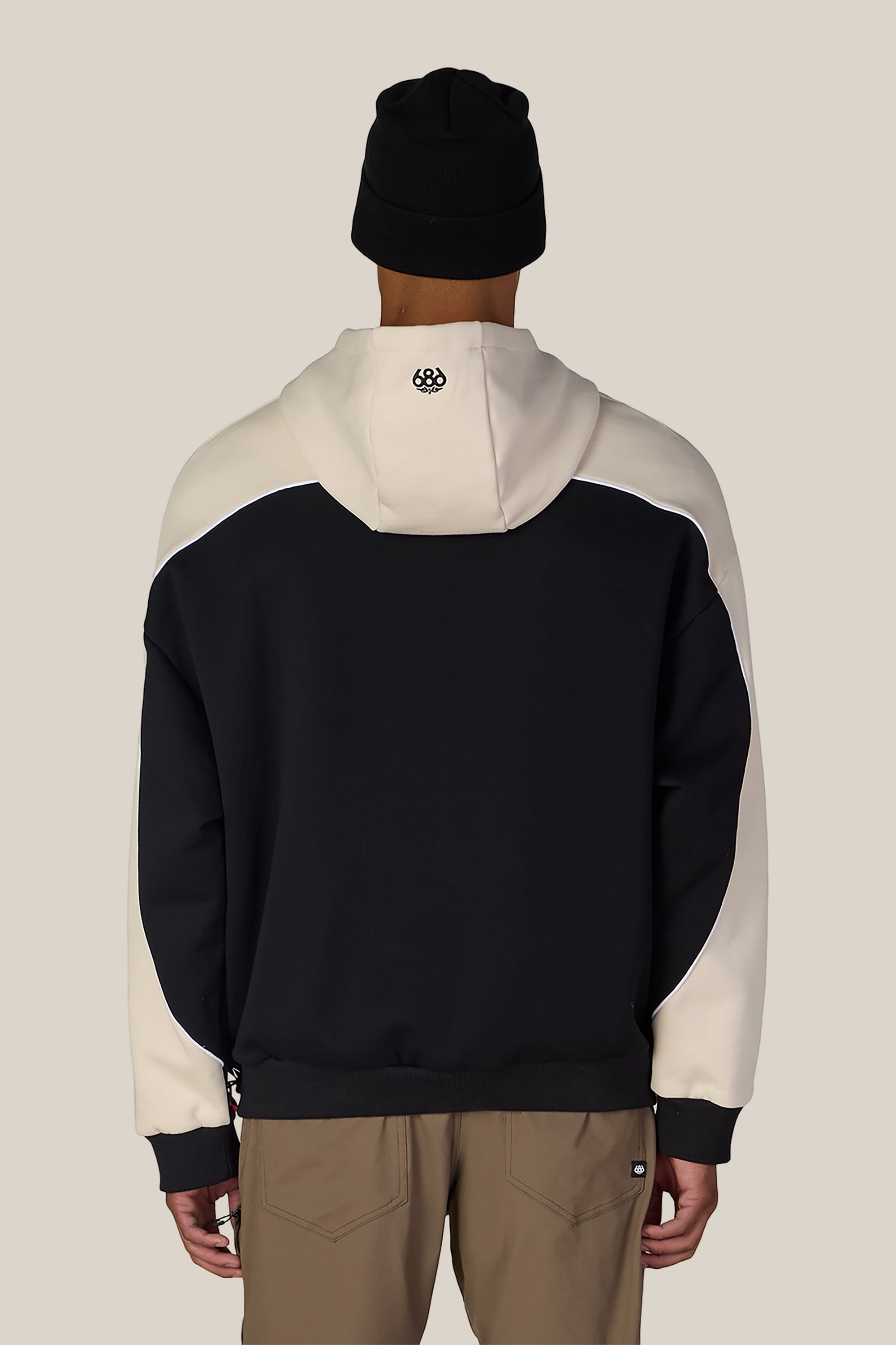 A person wearing a black beanie, brown pants, and the 686 Legacy Premium Heavyweight Hoody by 686—featuring a black and cream color scheme with a small logo on the hood—stands facing away from the camera against a plain background.