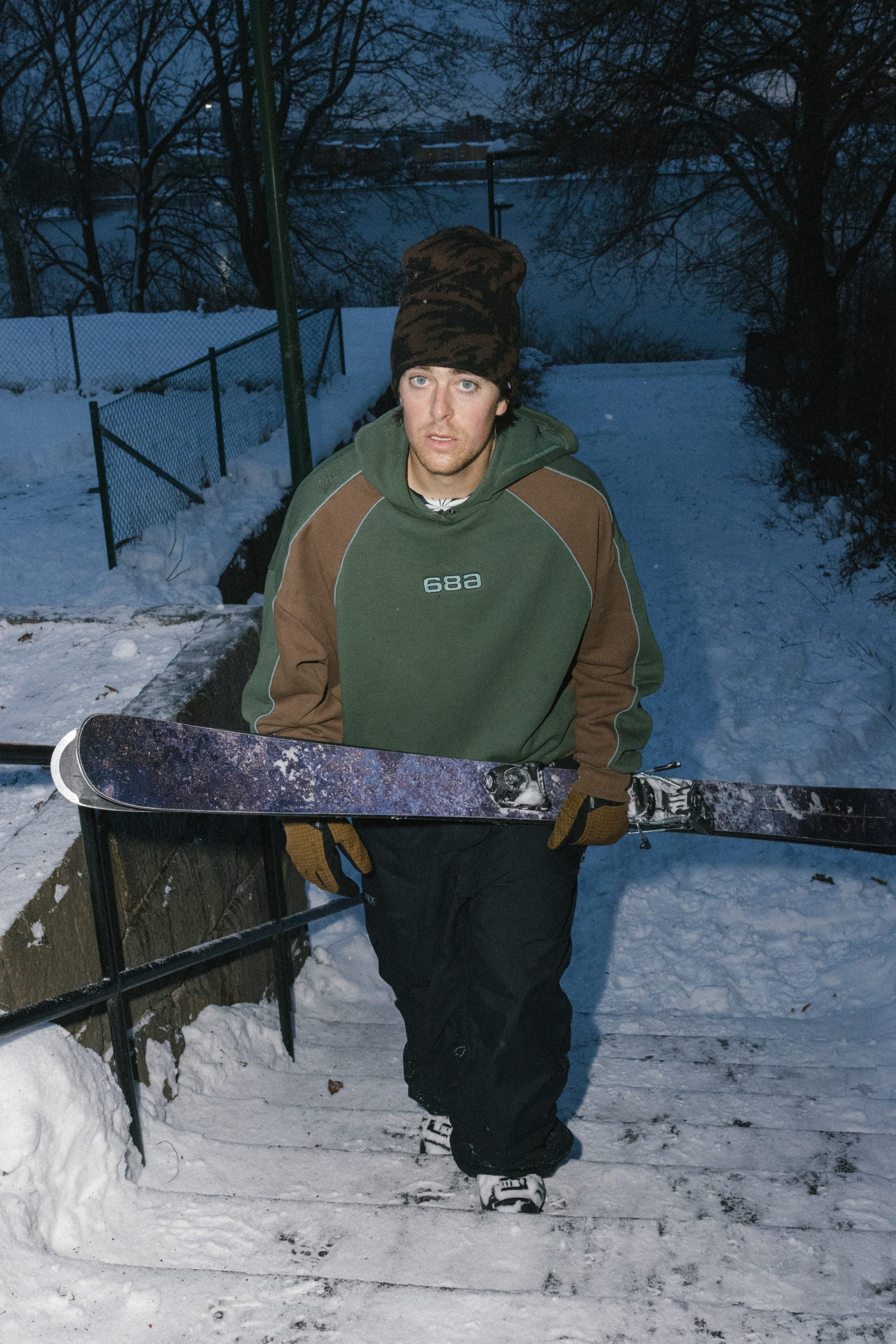 Wearing the 686 Legacy Premium Heavyweight Hoody from 686, a person in winter clothing holds a snowboard while walking up snowy outdoor stairs, with trees and a body of water in the background.