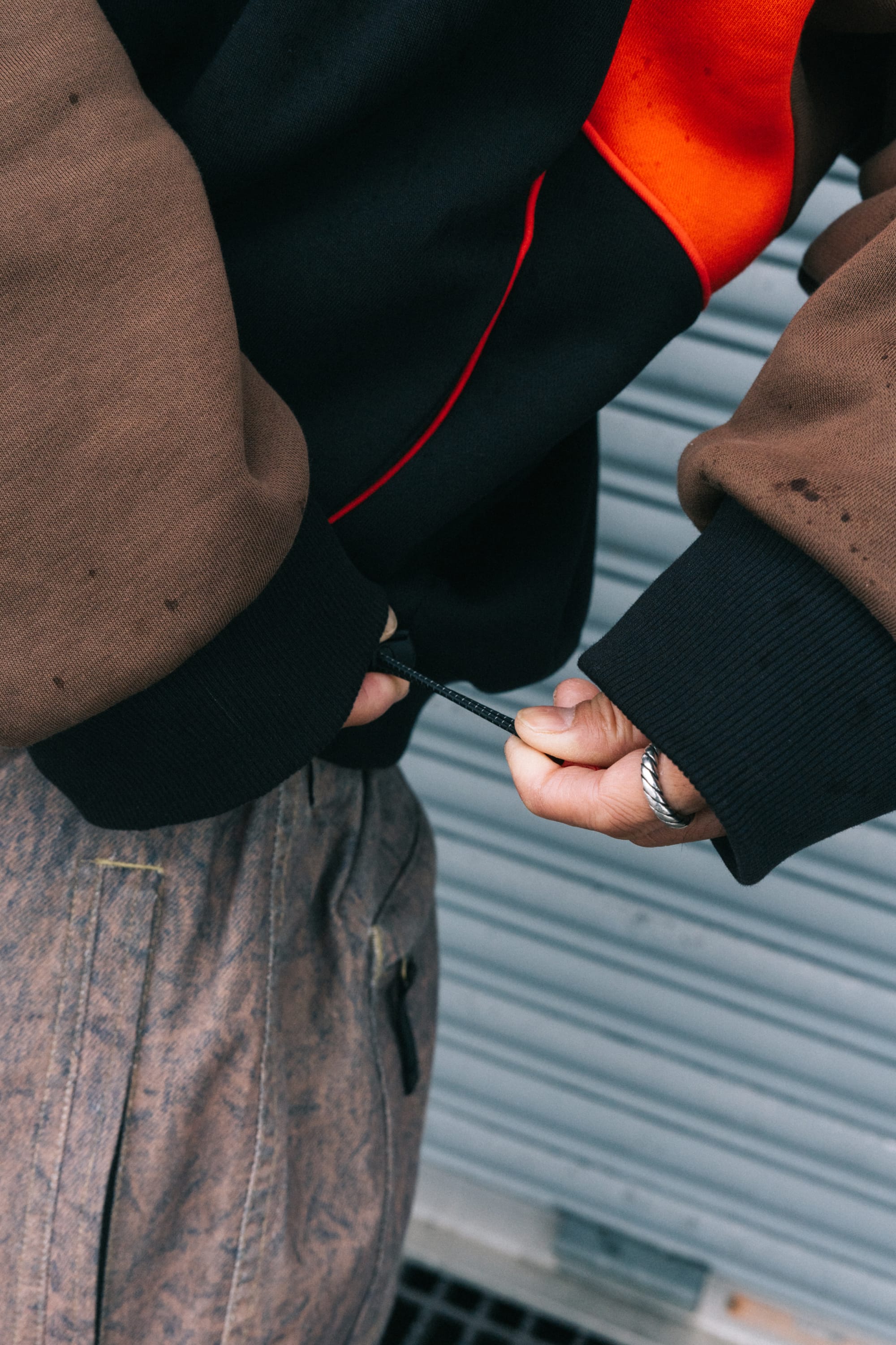 A person adjusts the drawstring on the bottom of a black and orange 686 Reserved® Premium Heavyweight Crewneck by 686, wearing brown pants with a zipper pocket and a silver ring on their finger. A metal roller shutter is visible in the background.