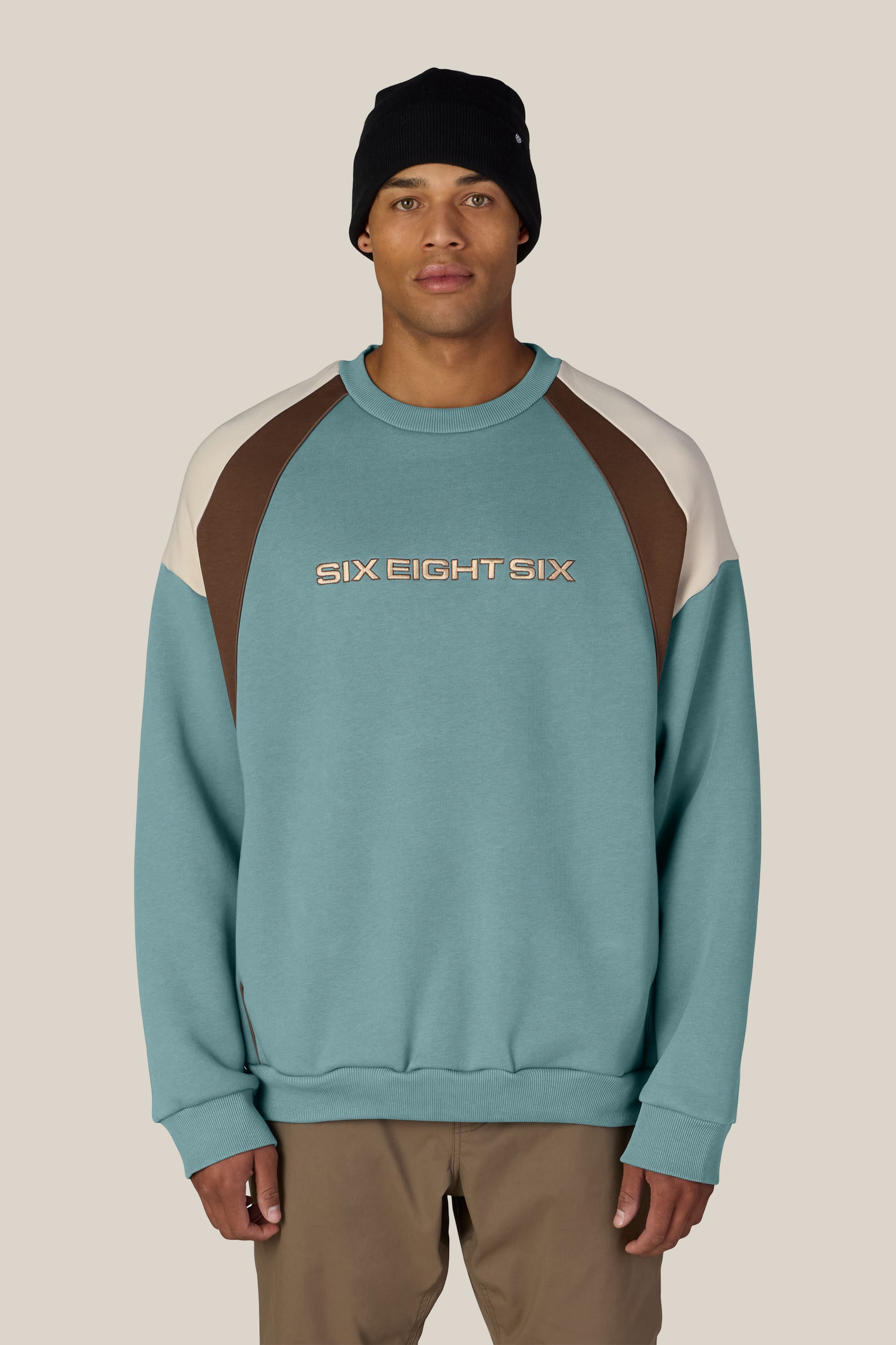 A person wearing a black beanie and the 686 Reserved® Premium Heavyweight Crewneck, a teal men’s cotton sweatshirt with brown and beige accents and SIX EIGHT SIX printed across the chest. The background is plain beige.