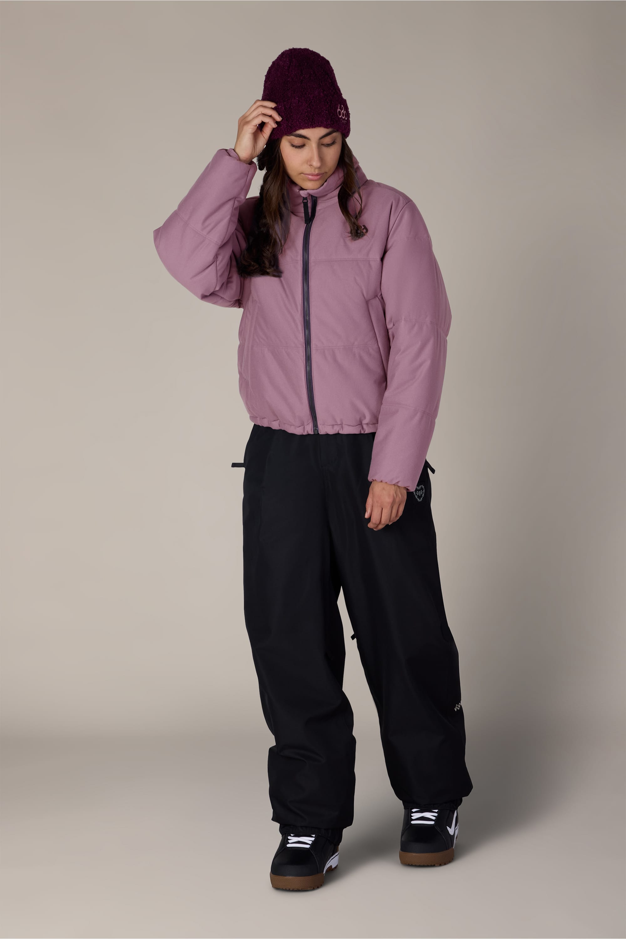 A person wearing the 686 Women’s Gracious Puffer Jacket in purple, paired with black pants, black shoes, and a maroon knit hat stands against a plain background, looking down while touching their hat.
