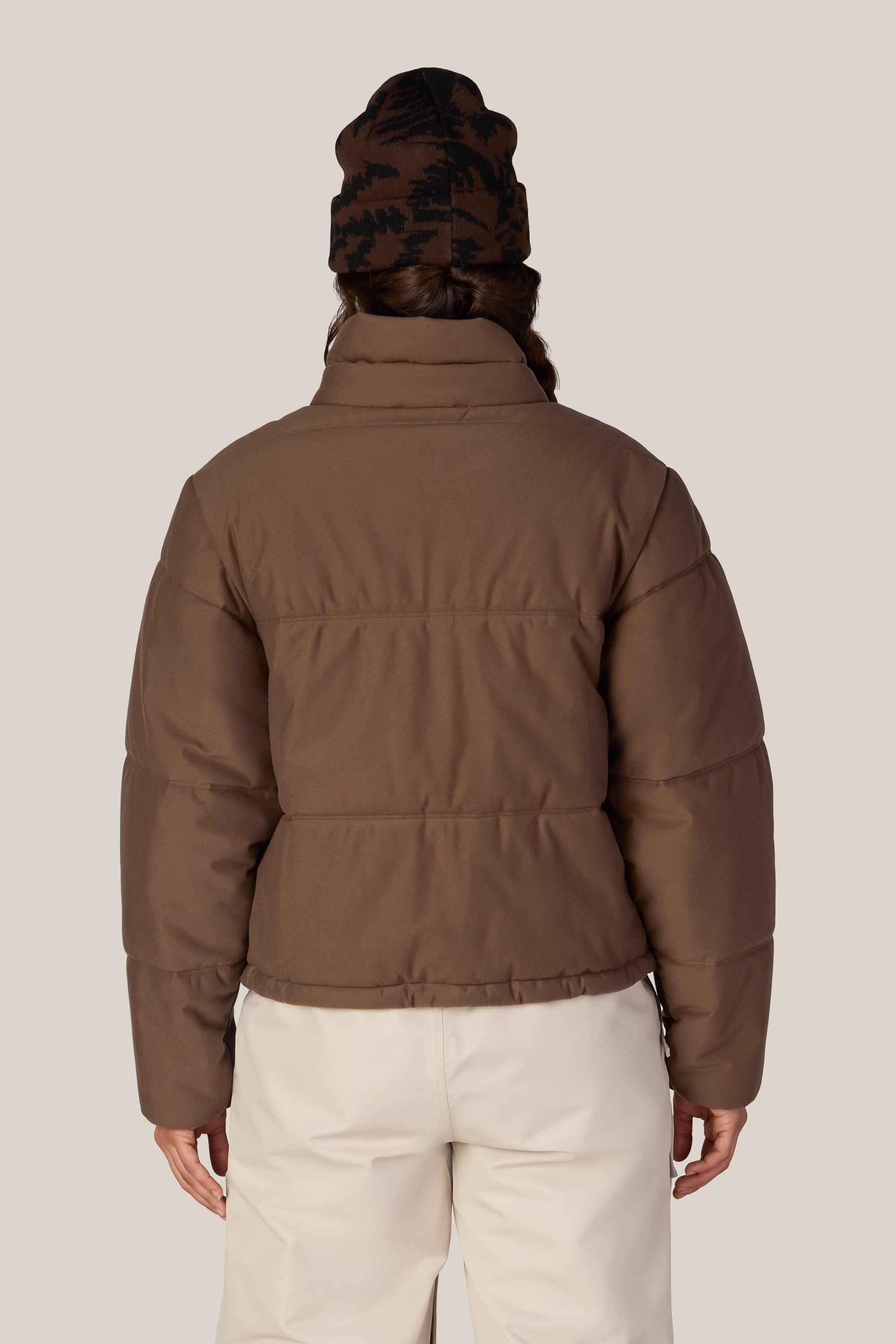 Person wearing the 686 Women’s Gracious Puffer Jacket in brown, paired with cream pants and a dark brown knit beanie, shown from the back against a plain light background.
