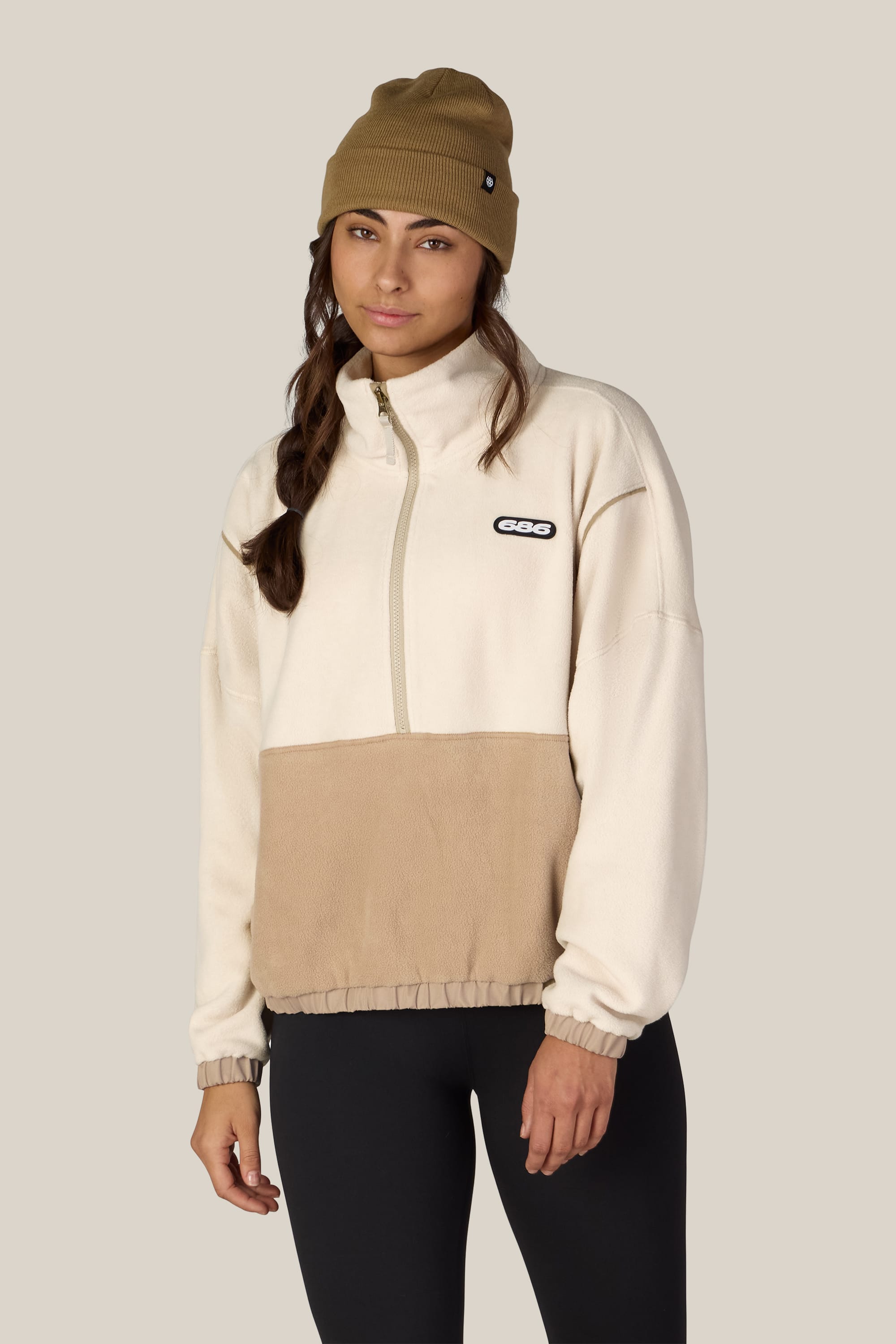 A woman wearing a tan beanie, black leggings, and the 686 Women’s Chimay Fleece Anorak by 686—a beige and tan pullover jacket made from 100% polyester—stands against a plain light background, looking at the camera with a neutral expression.