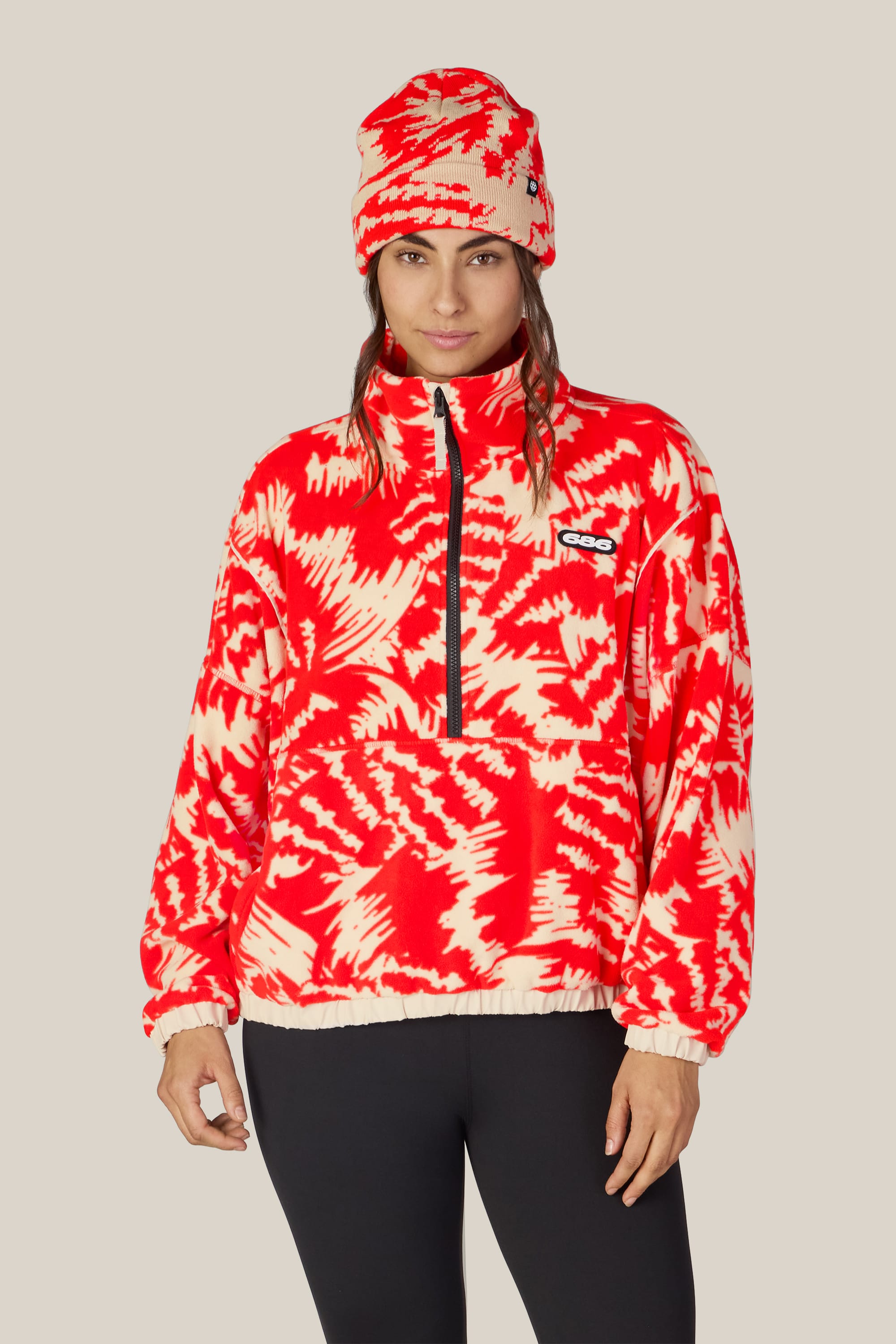 A person wearing the 686 Women’s Chimay Fleece Anorak—a red and white patterned, half-zip fleece pullover by 686—along with a matching beanie and black pants stands against a plain beige background, highlighting the cozy style of this women’s jacket.