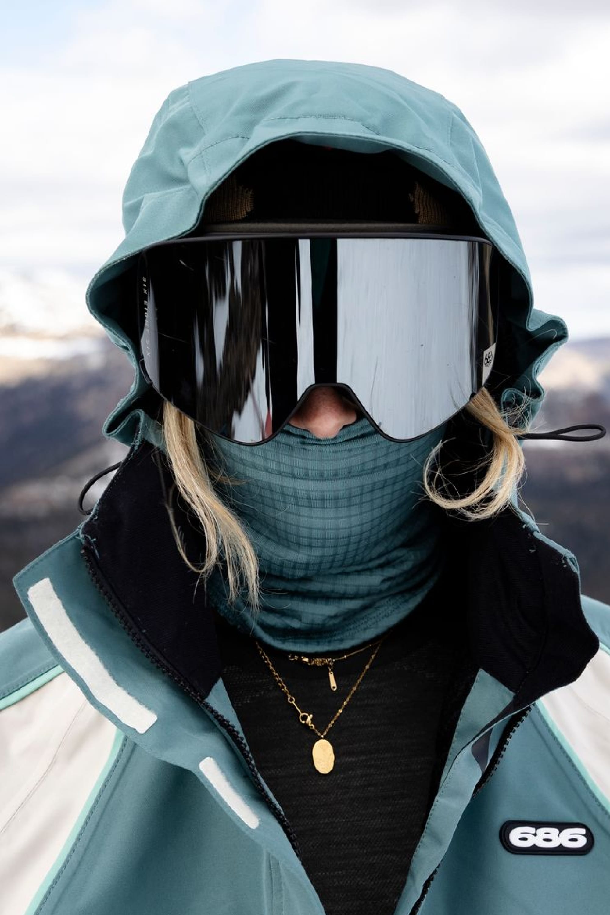 A person wearing a teal hooded jacket, large reflective ski goggles, and the 686 Mission Grid™ Balaclava by 686 stands outdoors with mountains and clouds in the background.