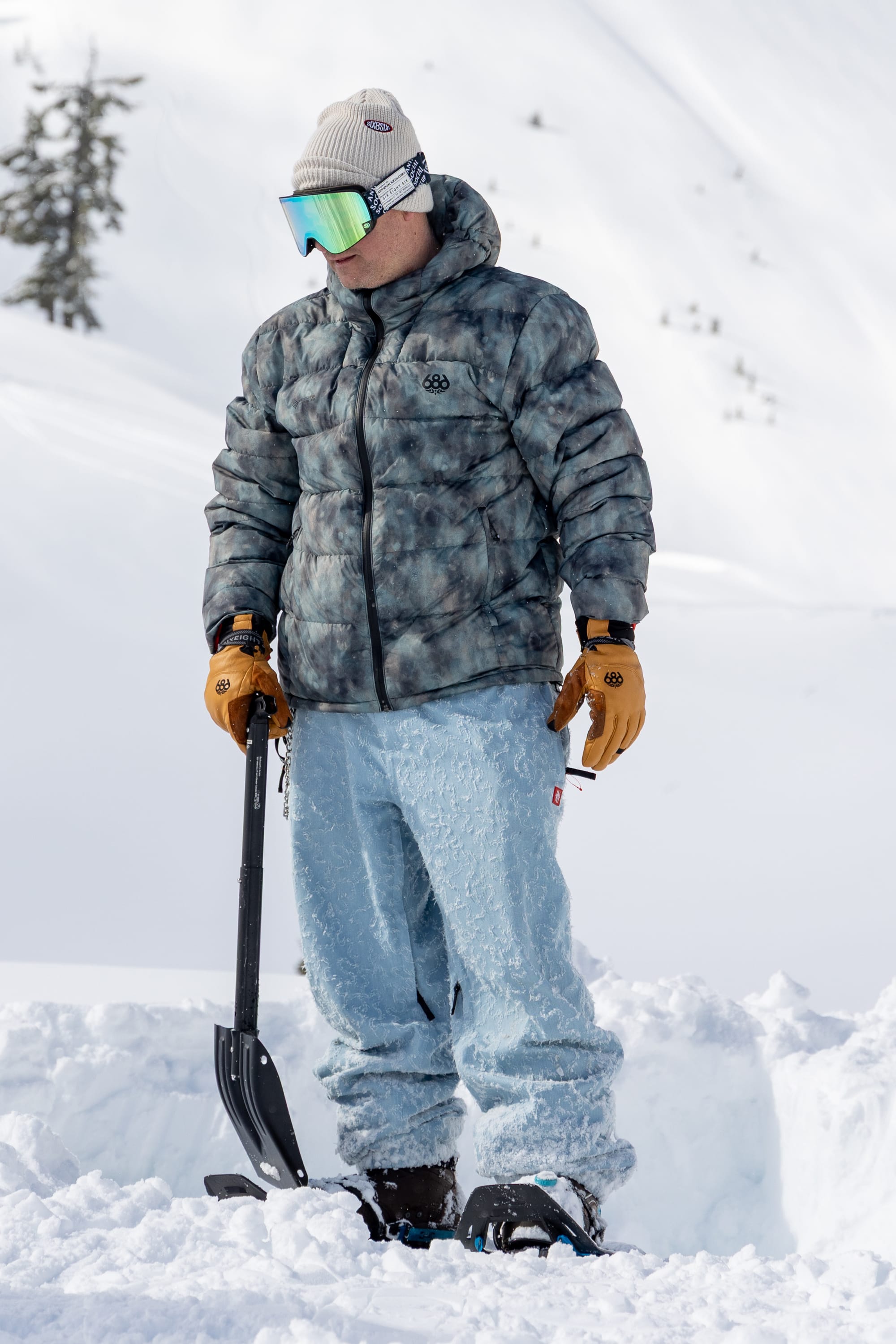 A person in a puffy jacket, snow pants, and the 686 Men’s GORE-TEX® Stronghold Leather Glove stands in the snow holding a shovel, with a snowy landscape and trees visible in the background.