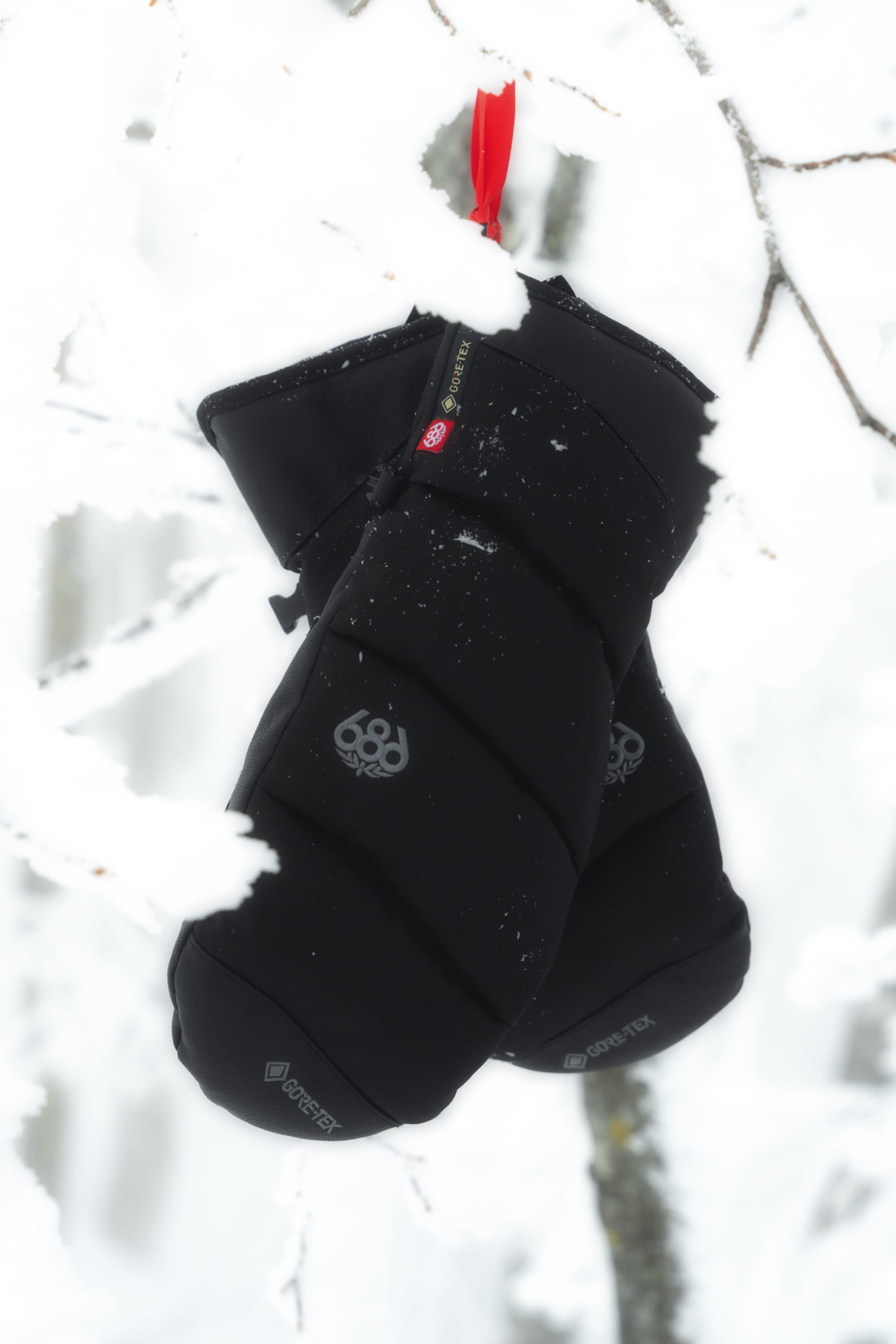 A pair of 686 Men’s GORE-TEX® Buffer Insulated Mitts in black, featuring a red loop, are hanging on a snow-covered tree branch in a snowy outdoor setting.