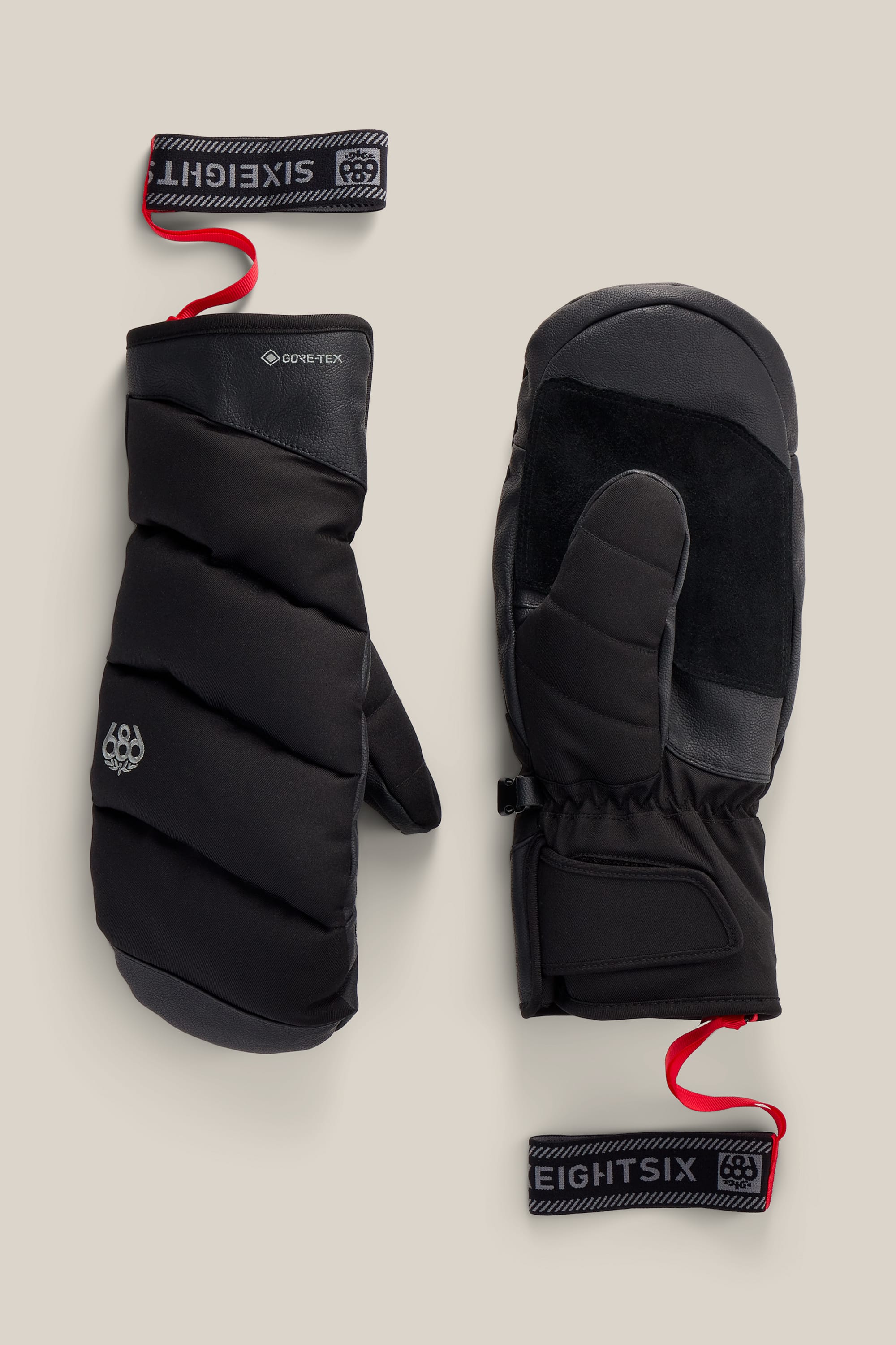 A pair of 686 Men’s GORE-TEX® Buffer Insulated Mitts in black, featuring PU construction and wrist straps, are displayed on a beige background—one positioned palm side up showing grip material, the other showcasing the back with a logo and GORE-TEX branding.
