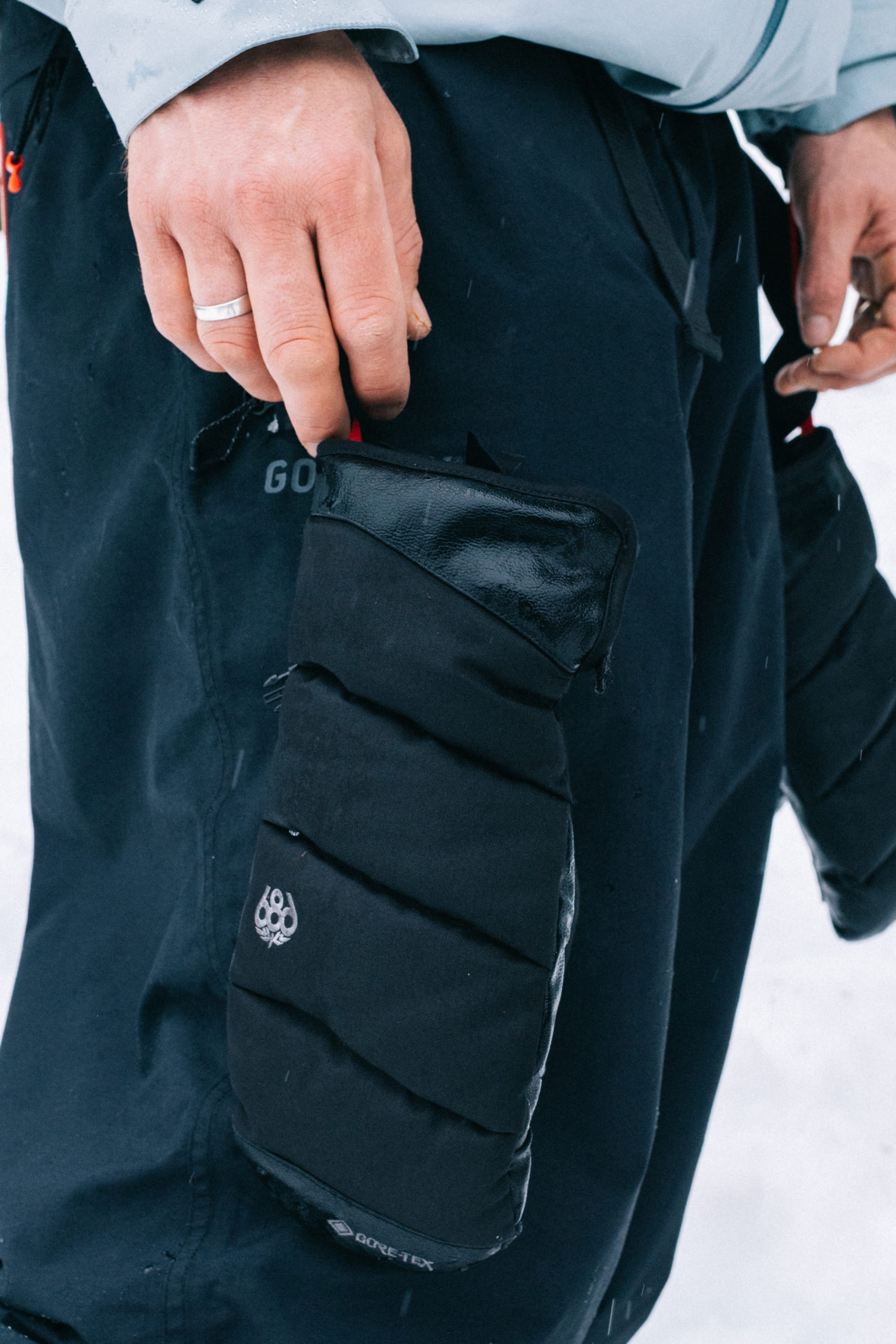 A close-up of a person’s gloved hand, wearing the sleek 686 Men’s GORE-TEX® Buffer Insulated Mitt, resting on black insulated pants with a zip pocket. Snow is visible in the background, and the person is wearing a silver ring on their finger.