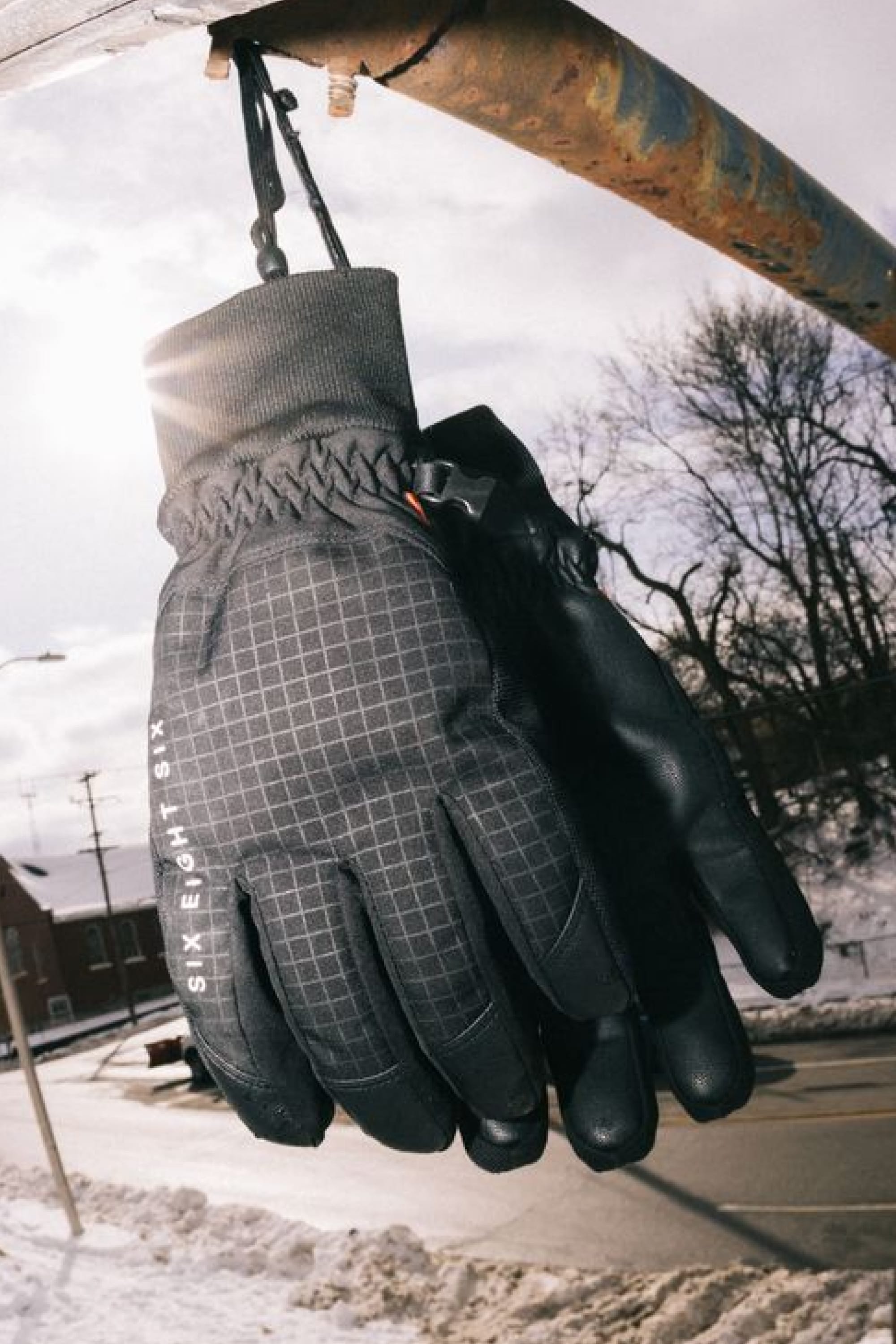 A pair of 686 Men’s Crush Insulated Gloves by 686 hang from a metal bar outdoors on a snowy day, with sunlight and bare trees in the background.