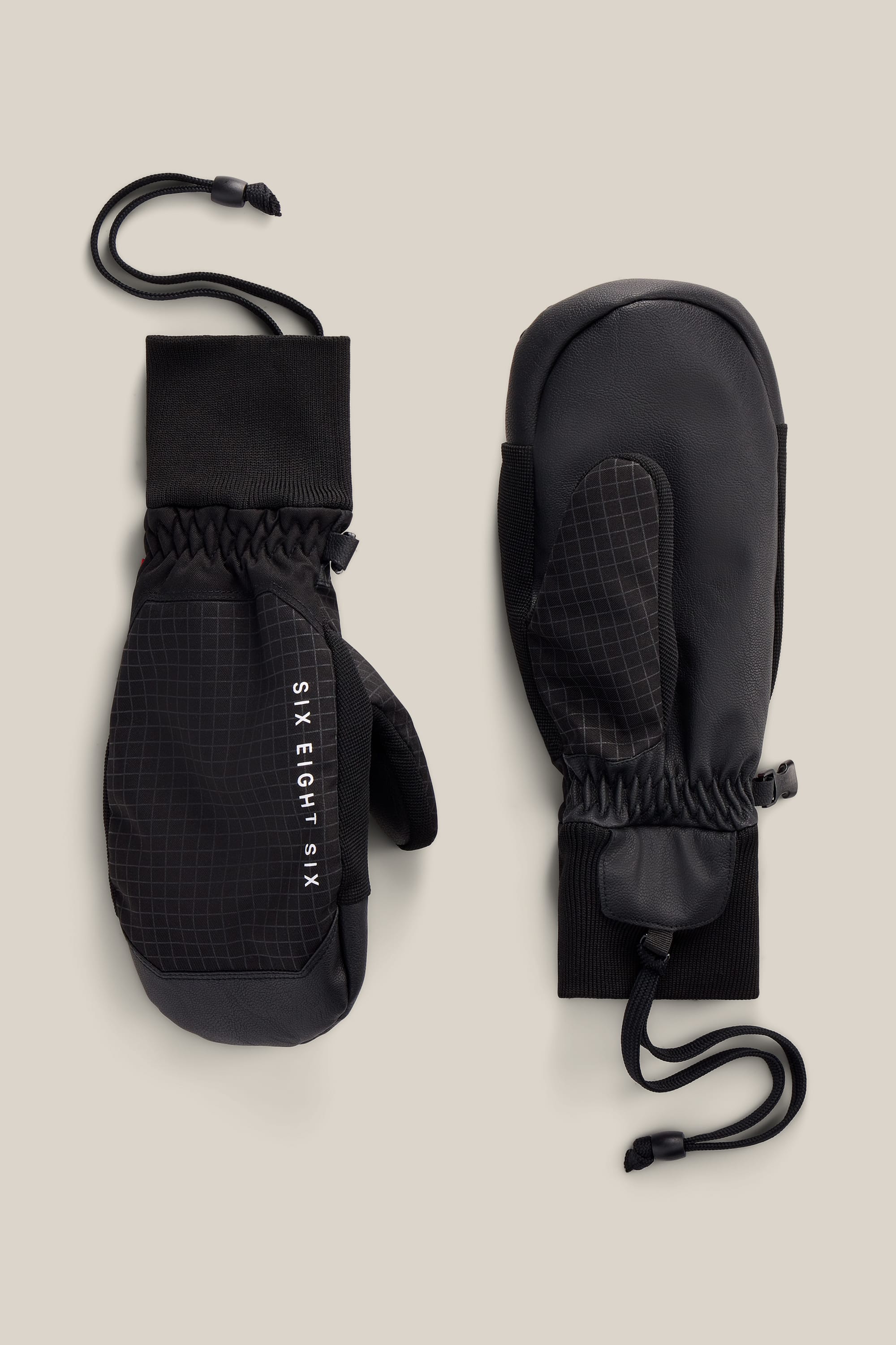 A pair of 686 Men’s Crush Insulated Mitts in black, featuring a grid pattern on the top, adjustable wrist straps, elastic cuffs, and attached wrist cords. One mitten displays the 686 brand name.