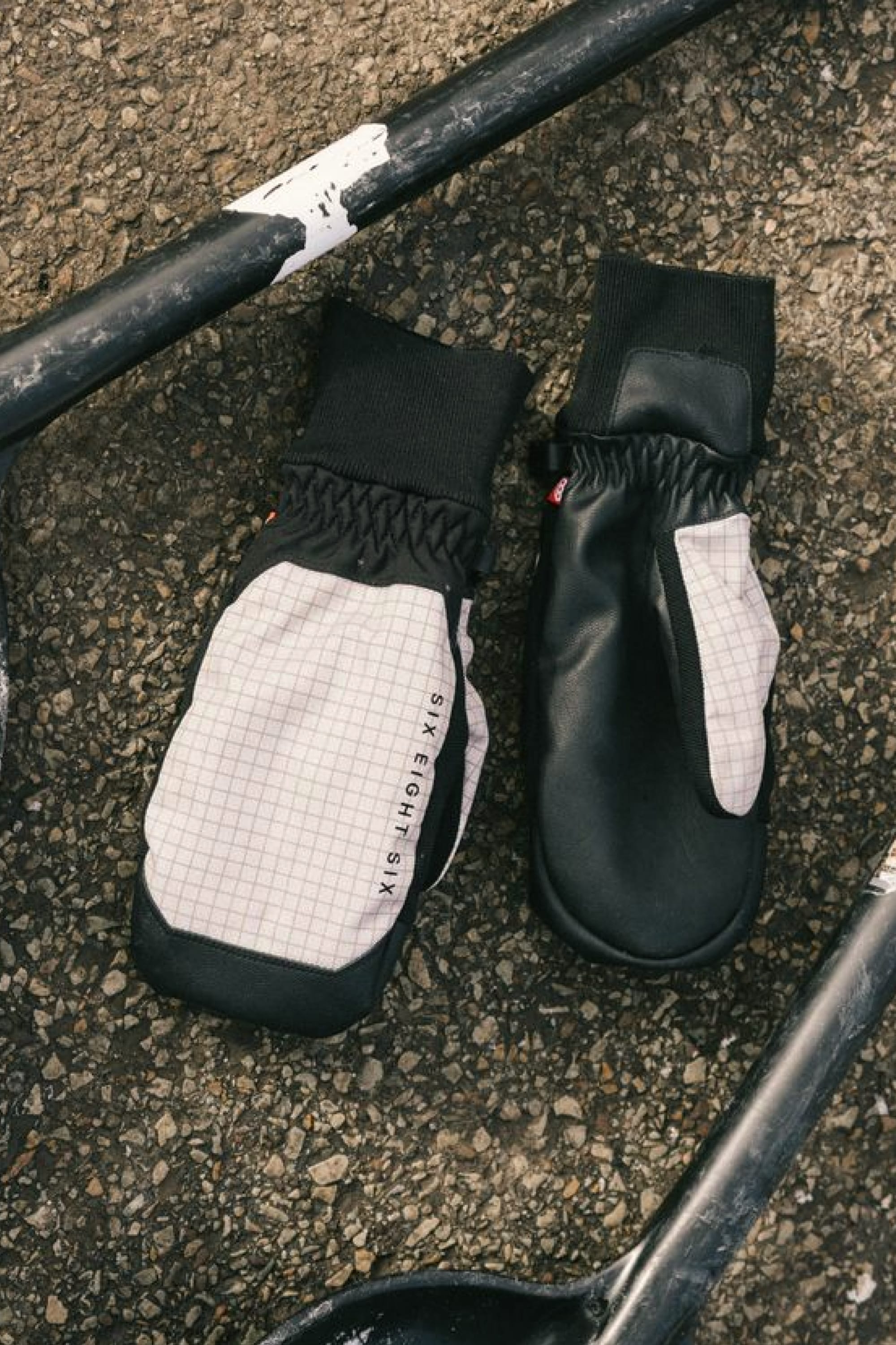 A pair of 686 Men’s Crush Insulated Mitts from 686, featuring a black and white grid pattern, lies on rough pavement next to black metal bars.