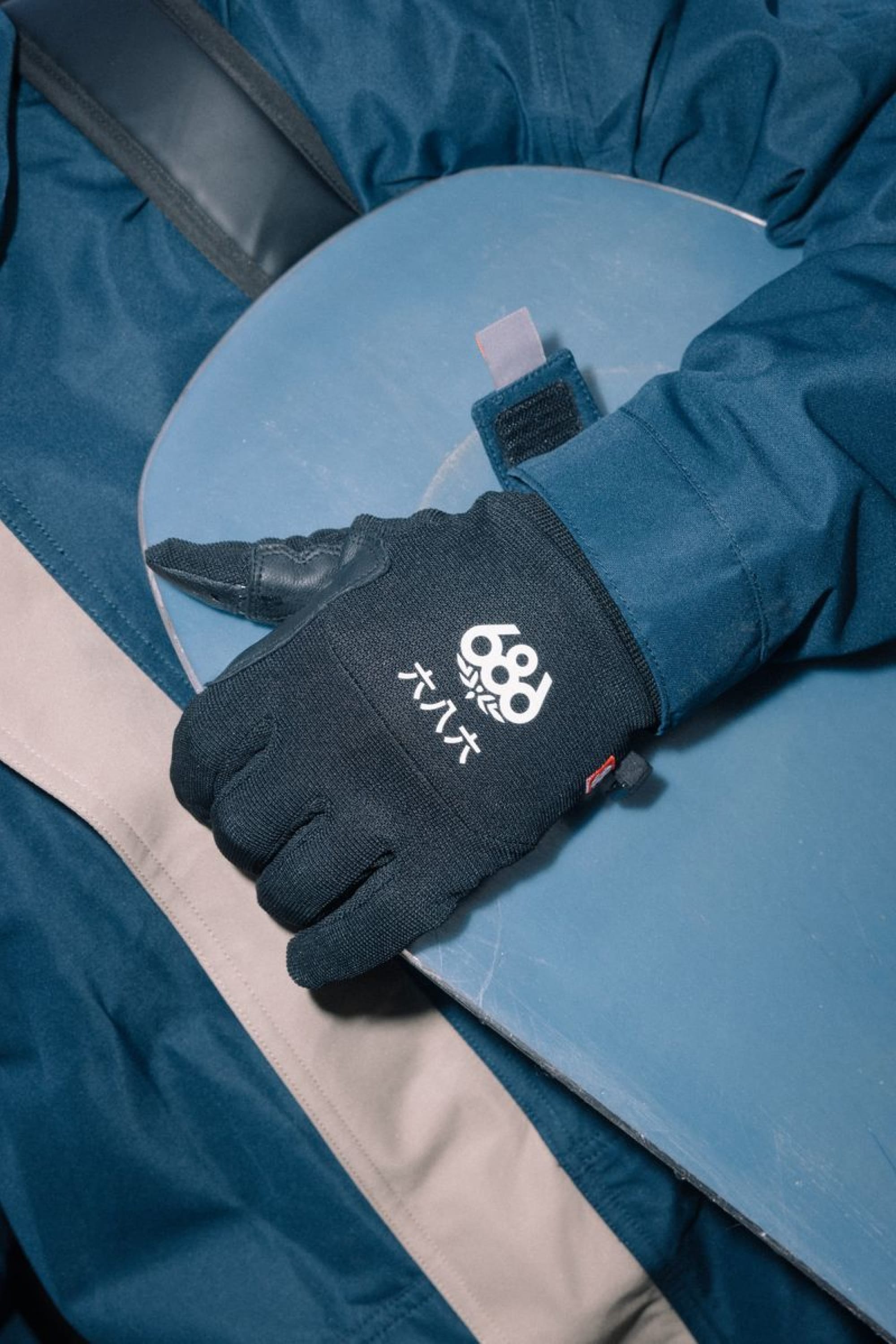 A person wearing a blue jacket and a pair of 686 Men’s Hold Up Pipe Gloves in black, featuring white text and symbols, rests their gloved hand on a blue snowboard.