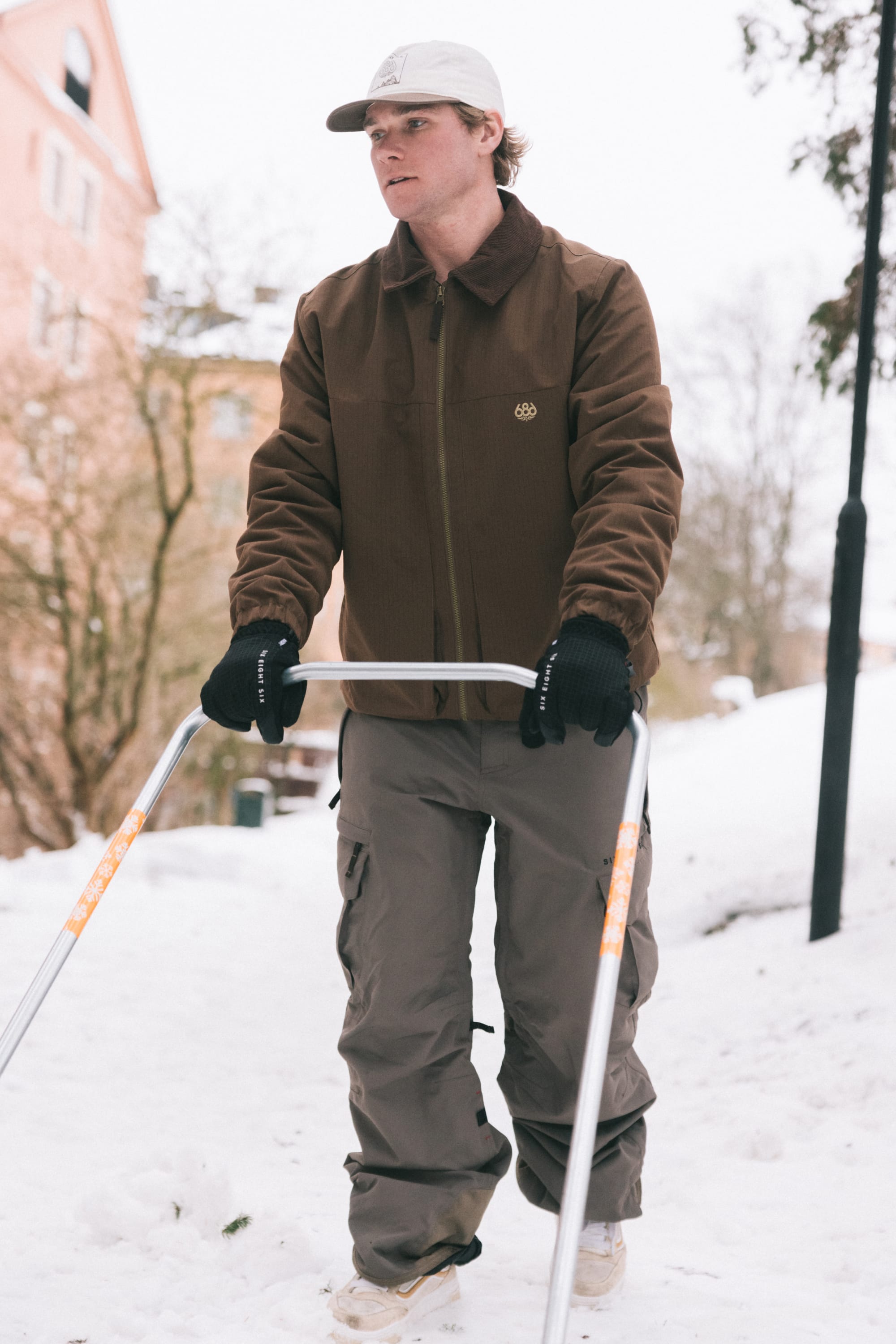 Wearing the 686 Men’s 686 Camber Insulated Jacket by 686 and gray pants, a person uses a walker on a snowy path with trees and a peach-colored building visible in the background.