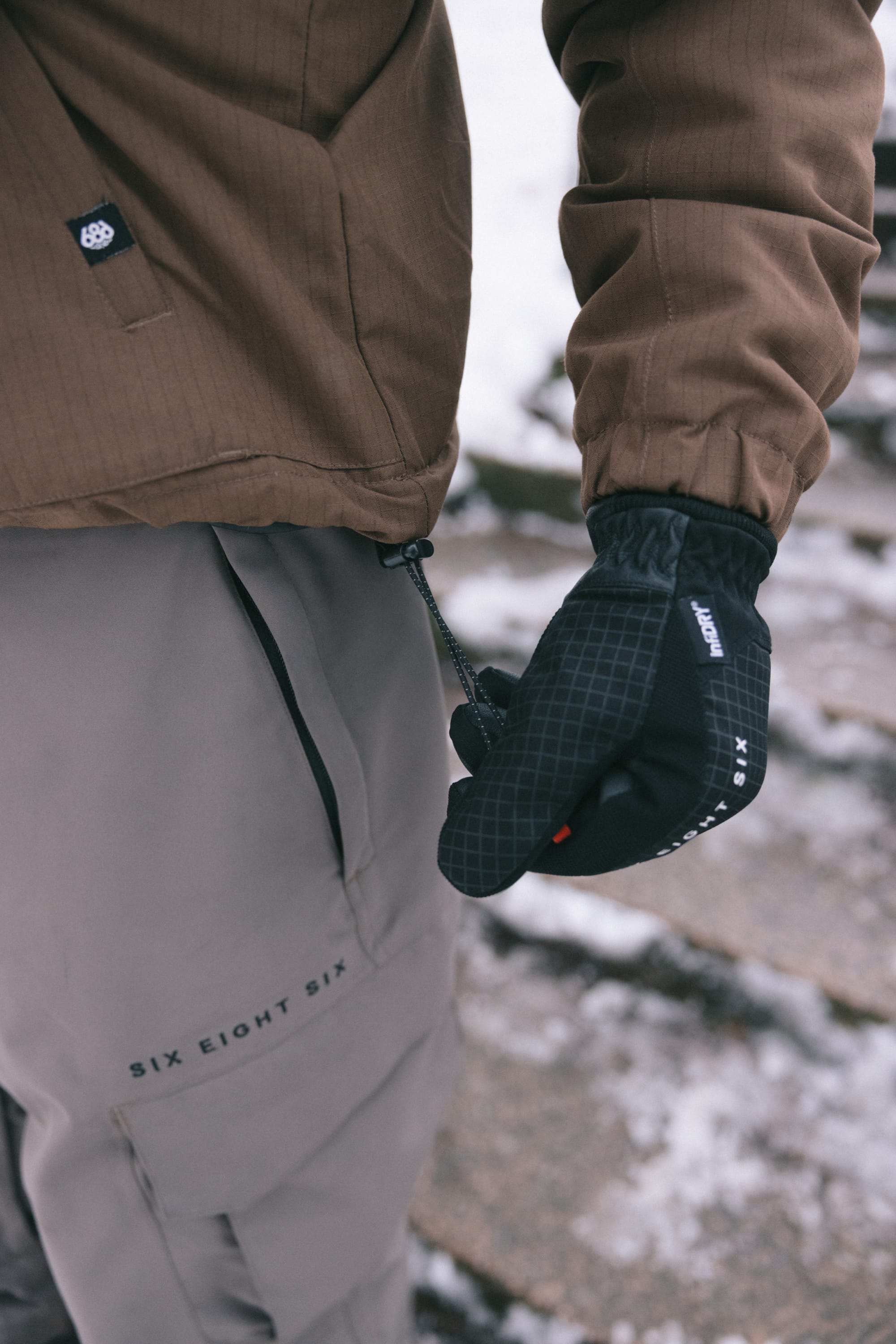 A person wearing the 686 Men’s 686 Camber Insulated Jacket—a brown men’s polyester jacket by 686—along with gray SIX EIGHT SIX pants and black gloves, stands outdoors on snowy stone steps, adjusting the jacket’s drawstring.