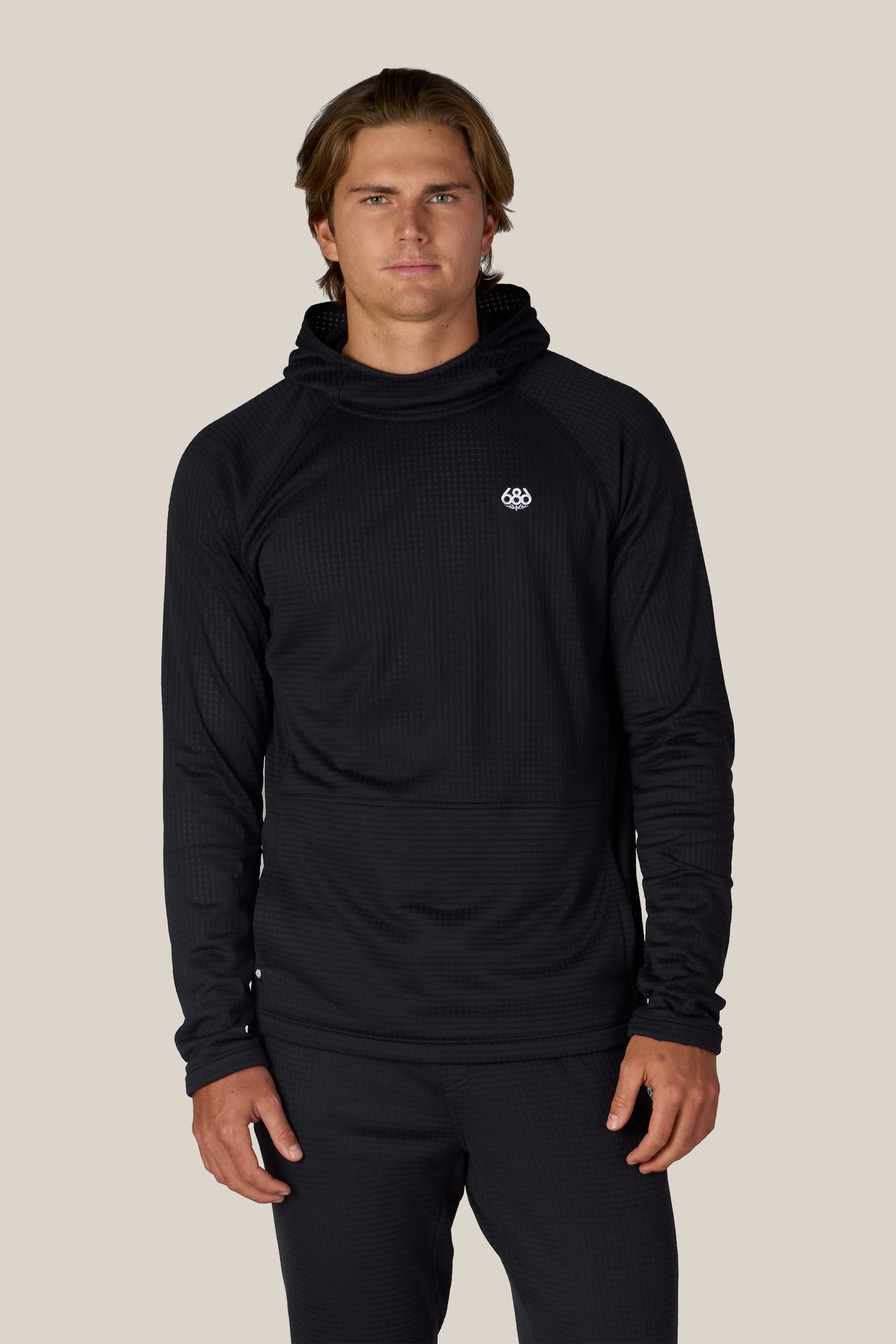 A man with light brown hair wears the 686 Men’s Mission Grid™ Raglan Hoody in black, standing against a plain beige background, facing forward with a neutral expression.