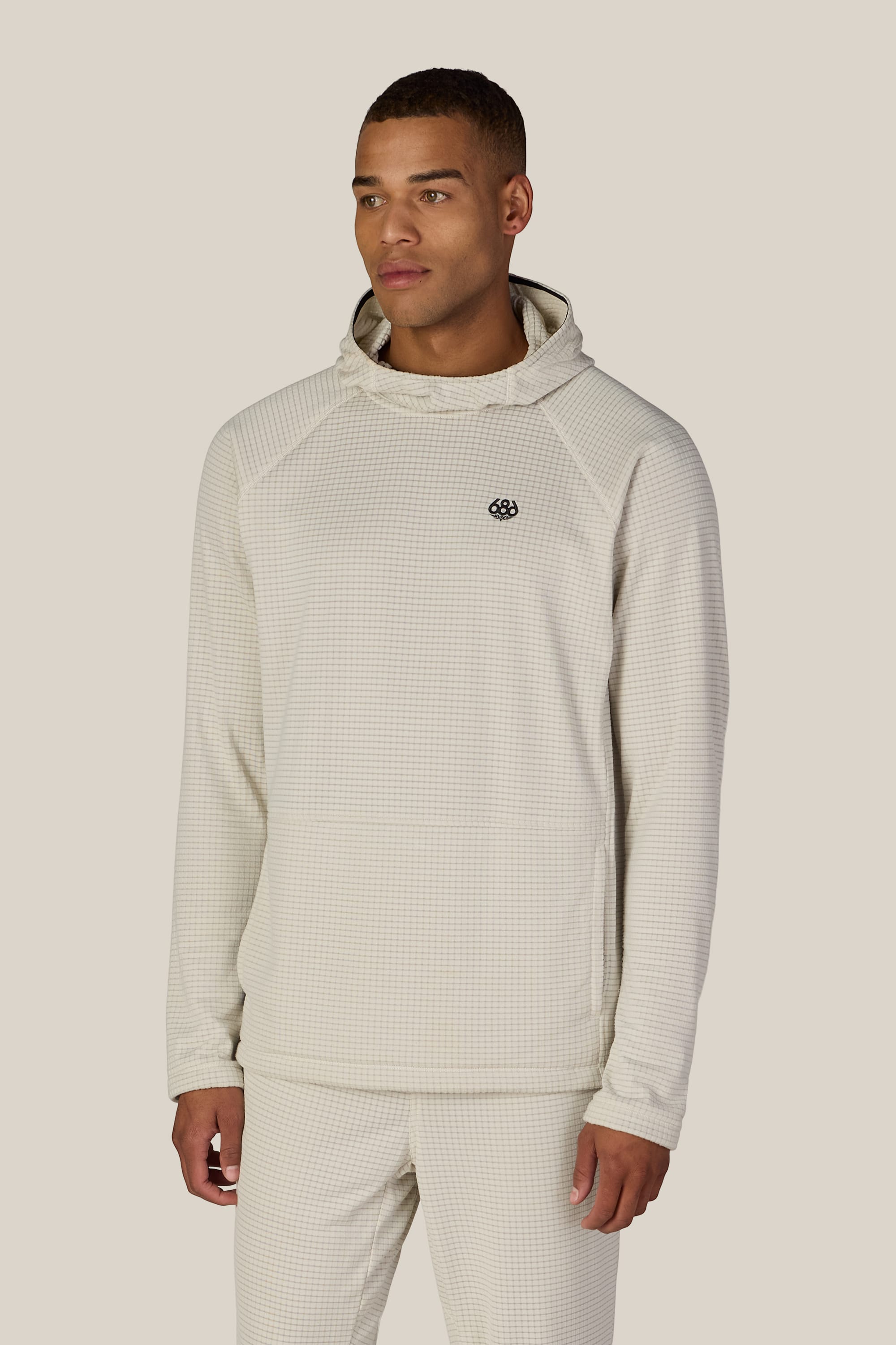 A man wearing the 686 Men’s Mission Grid™ Raglan Hoody in light beige stands facing slightly to the side against a plain beige background.