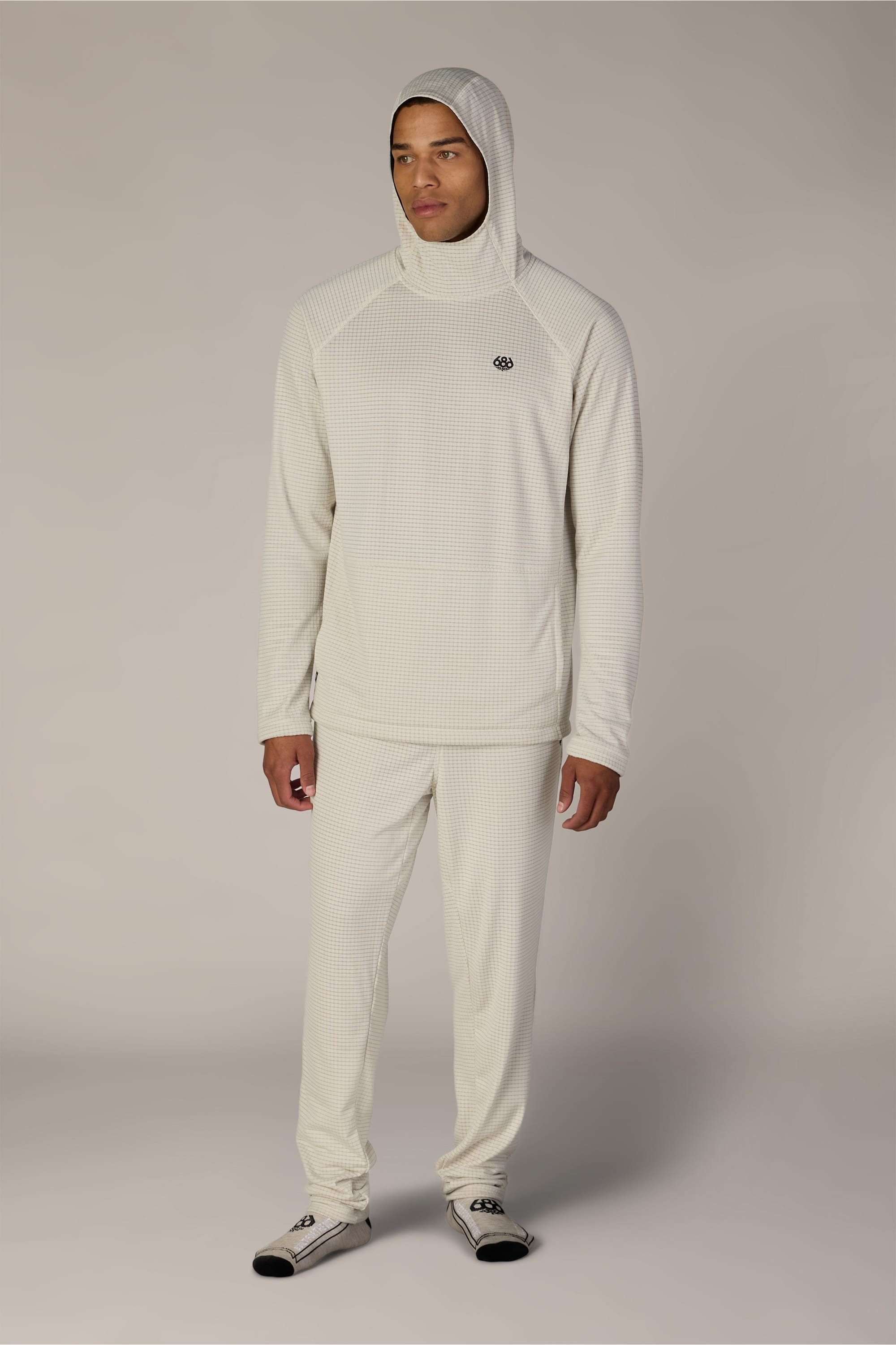 A man stands wearing the 686 Men’s Mission Grid™ Raglan Hoody by 686 and matching pants, featuring a logo on the chest, along with light socks. He faces slightly to the side against a plain, neutral background.